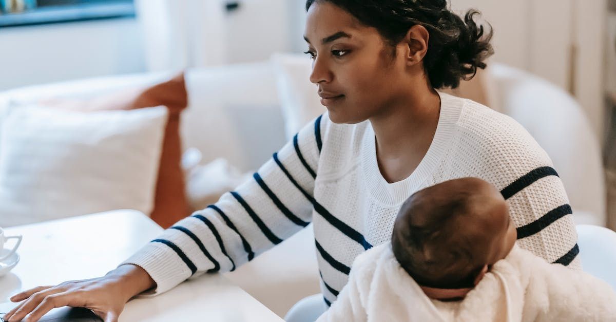 Woman with a baby on her lap works on a laptop in a bright living room. She wears a striped sweater, looking focused.