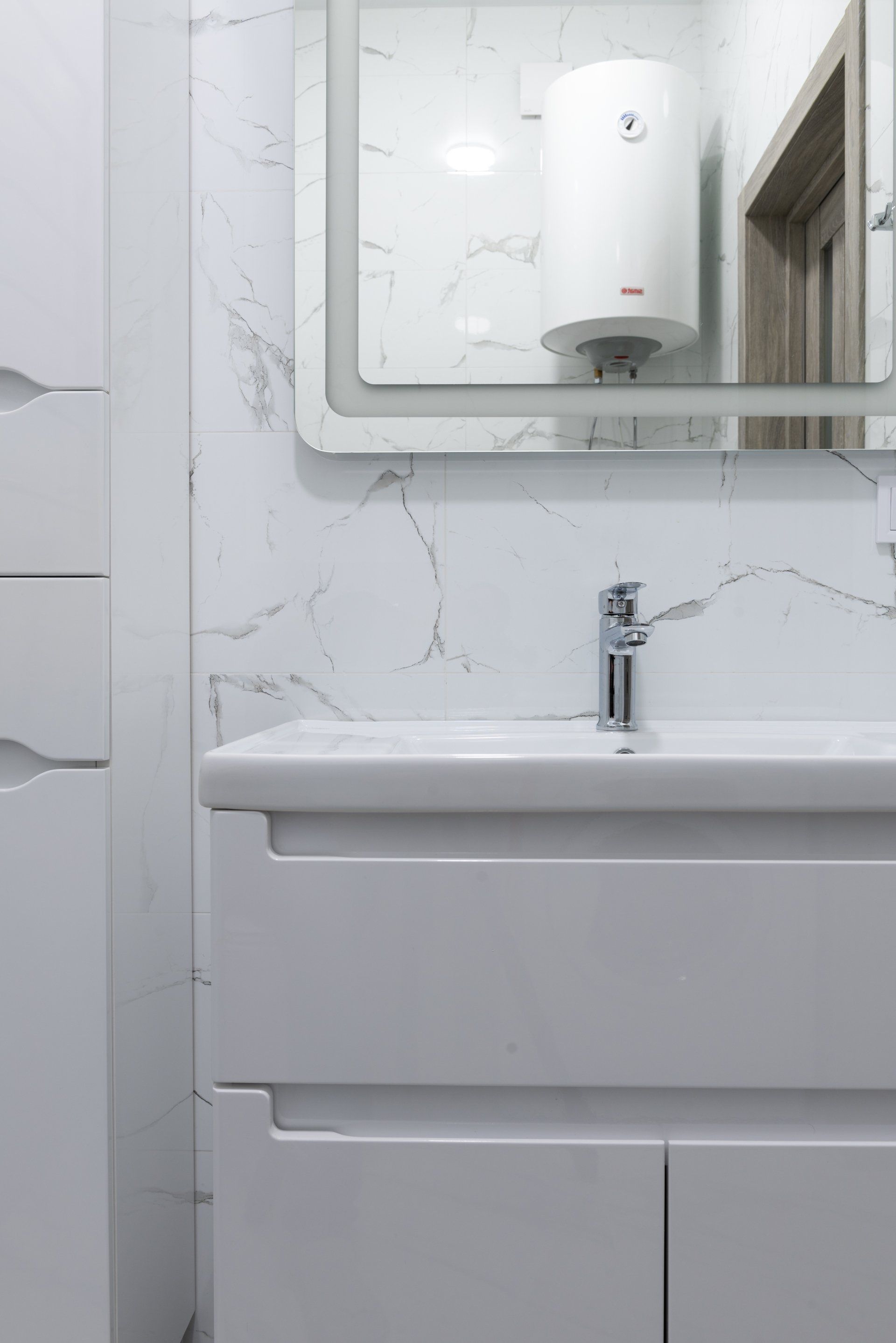 A modern bathroom sink vanity against a white marble-tiled wall with a water heater visible in the vanity mirror.