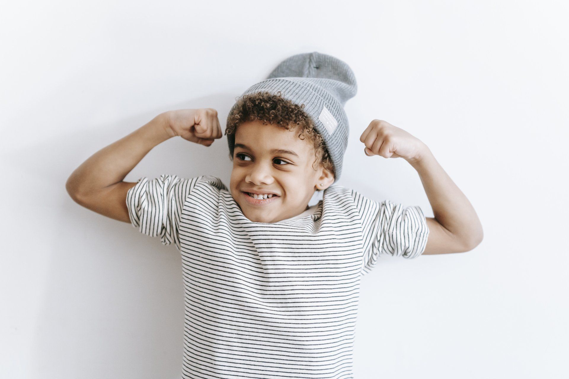 A child wearing a grey beanie and striped shirt flexes both arms against a plain white background.