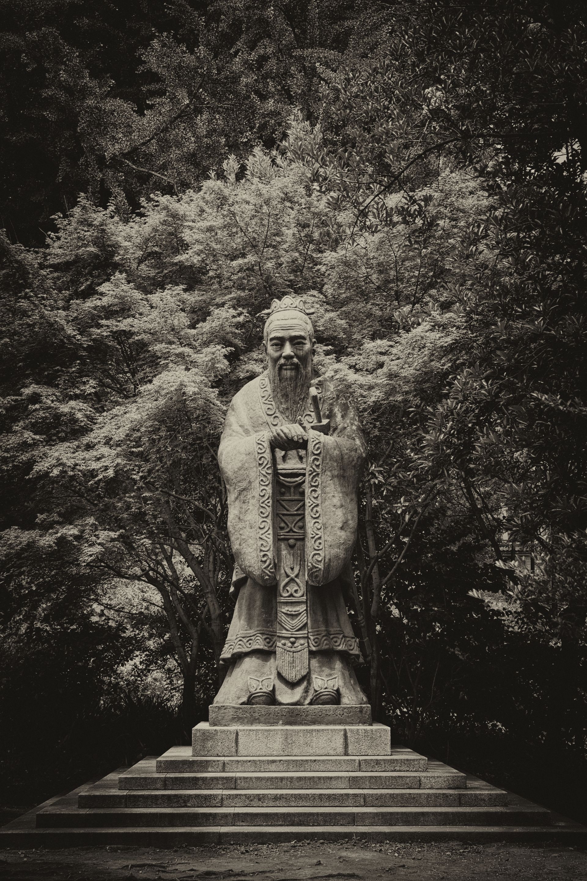 Statue of Confucius in traditional robes, standing in a park-like setting with lush trees in the background.