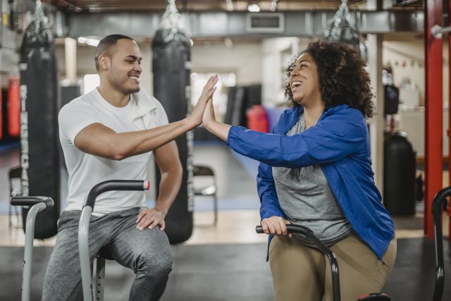 Deux personnes se tapent dans la main en étant assises sur des appareils de musculation dans une salle de sport.