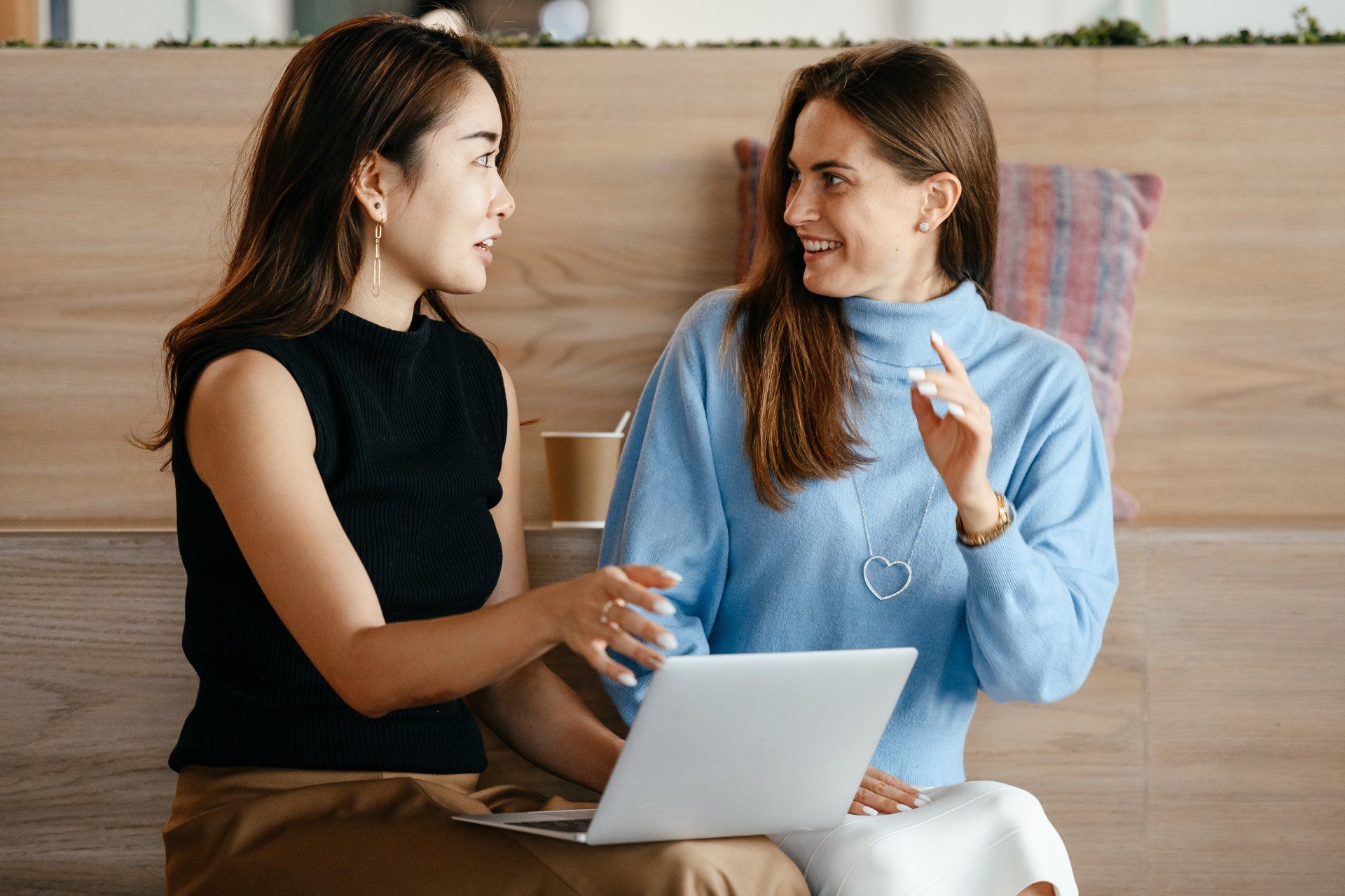 Two professional women are sitting  at a coffee shop on a bench with a laptop discussing their careers