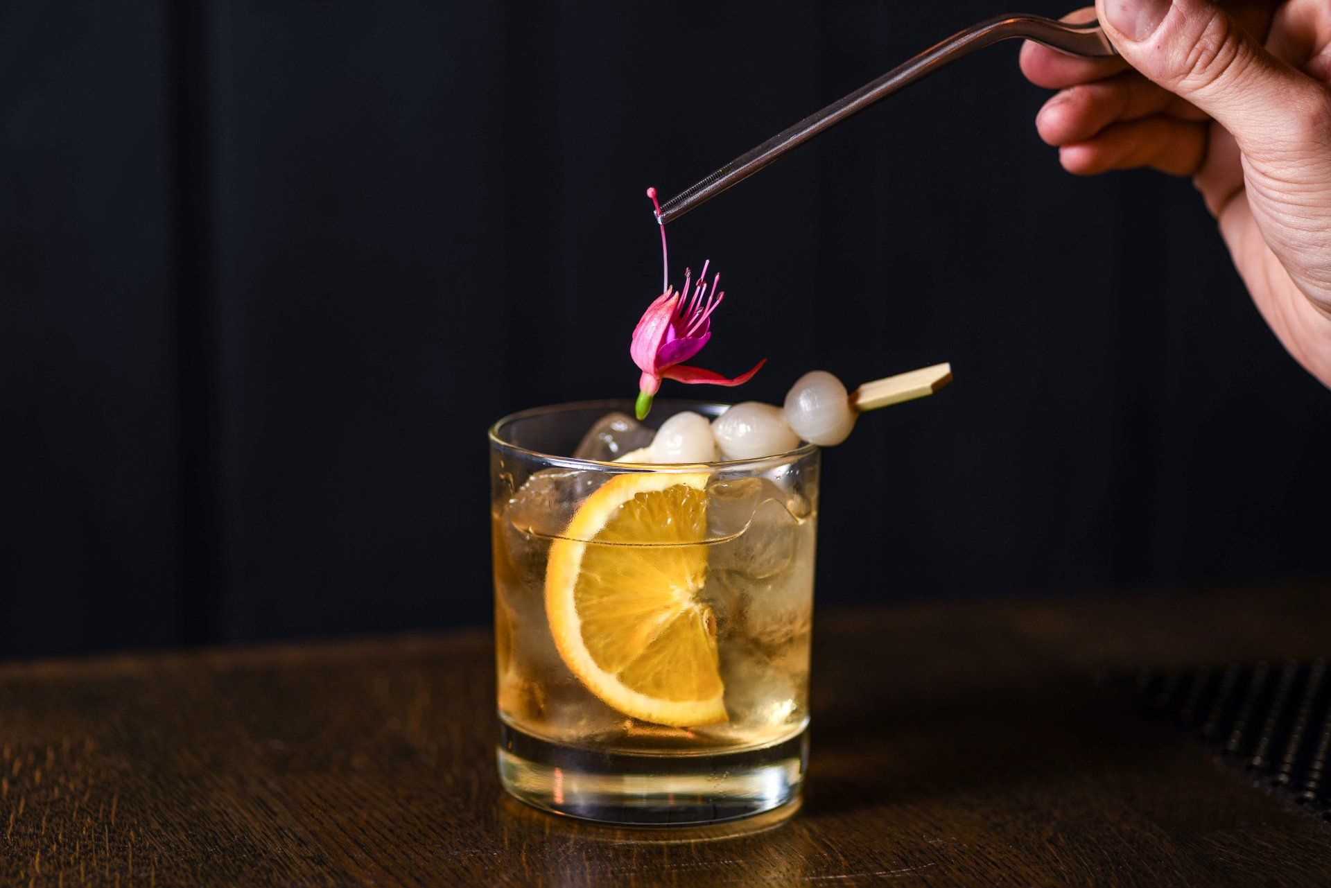 Cocktail with orange slice and skewered onions, topped with a flower, being garnished by hand.