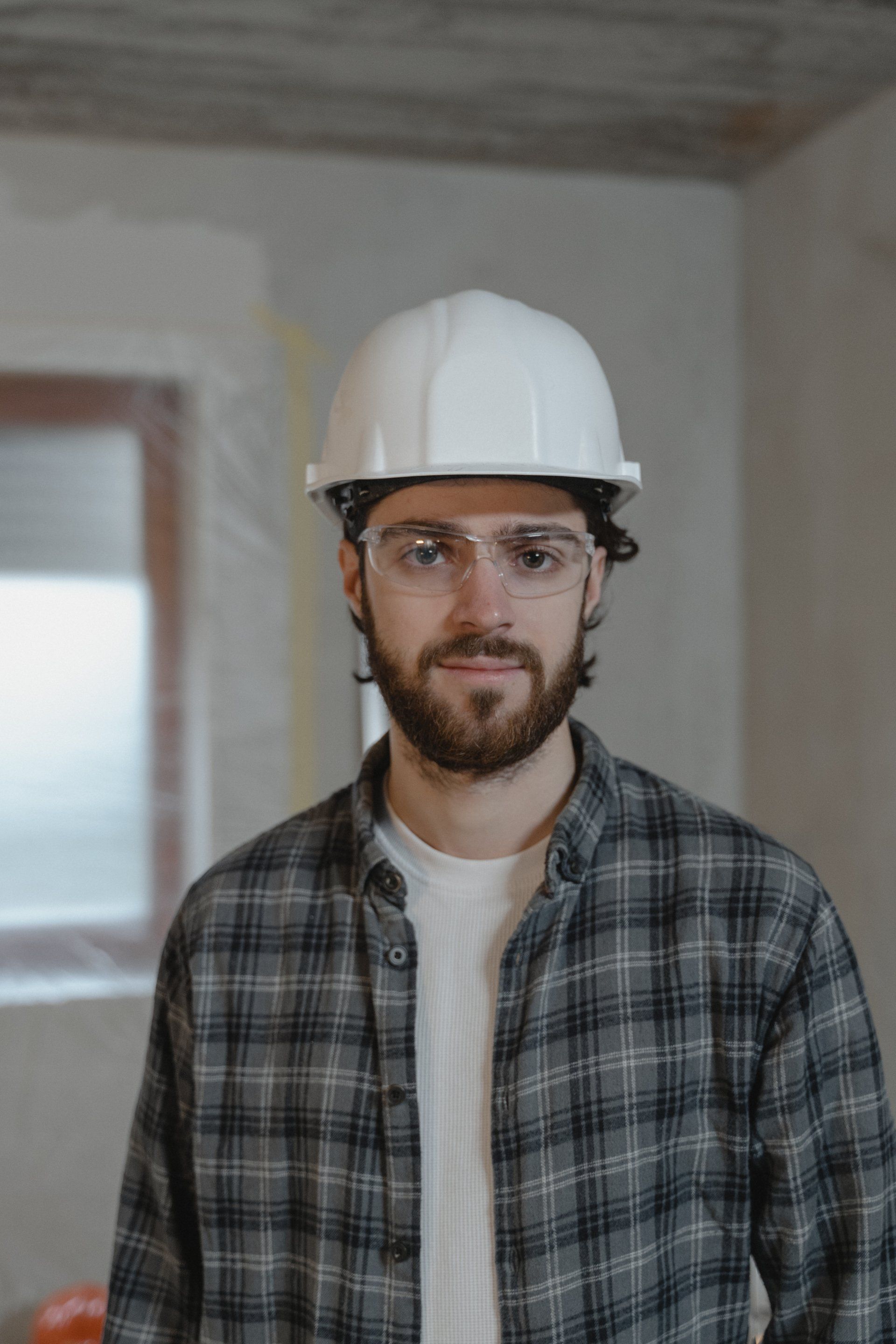 Construction worker in a white hard hat and glasses standing indoors at a renovation site.