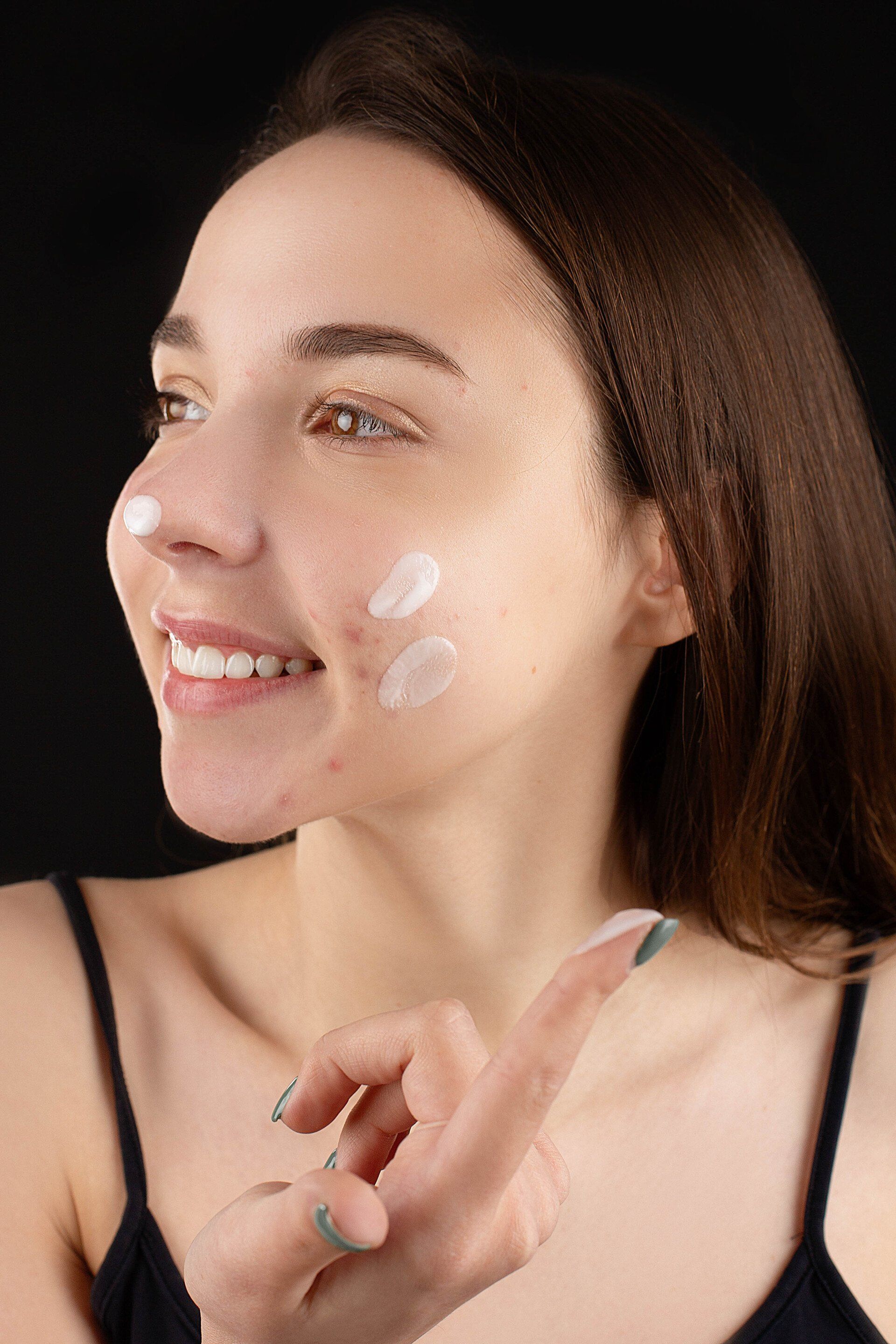 A woman is smiling while applying cream to her face.