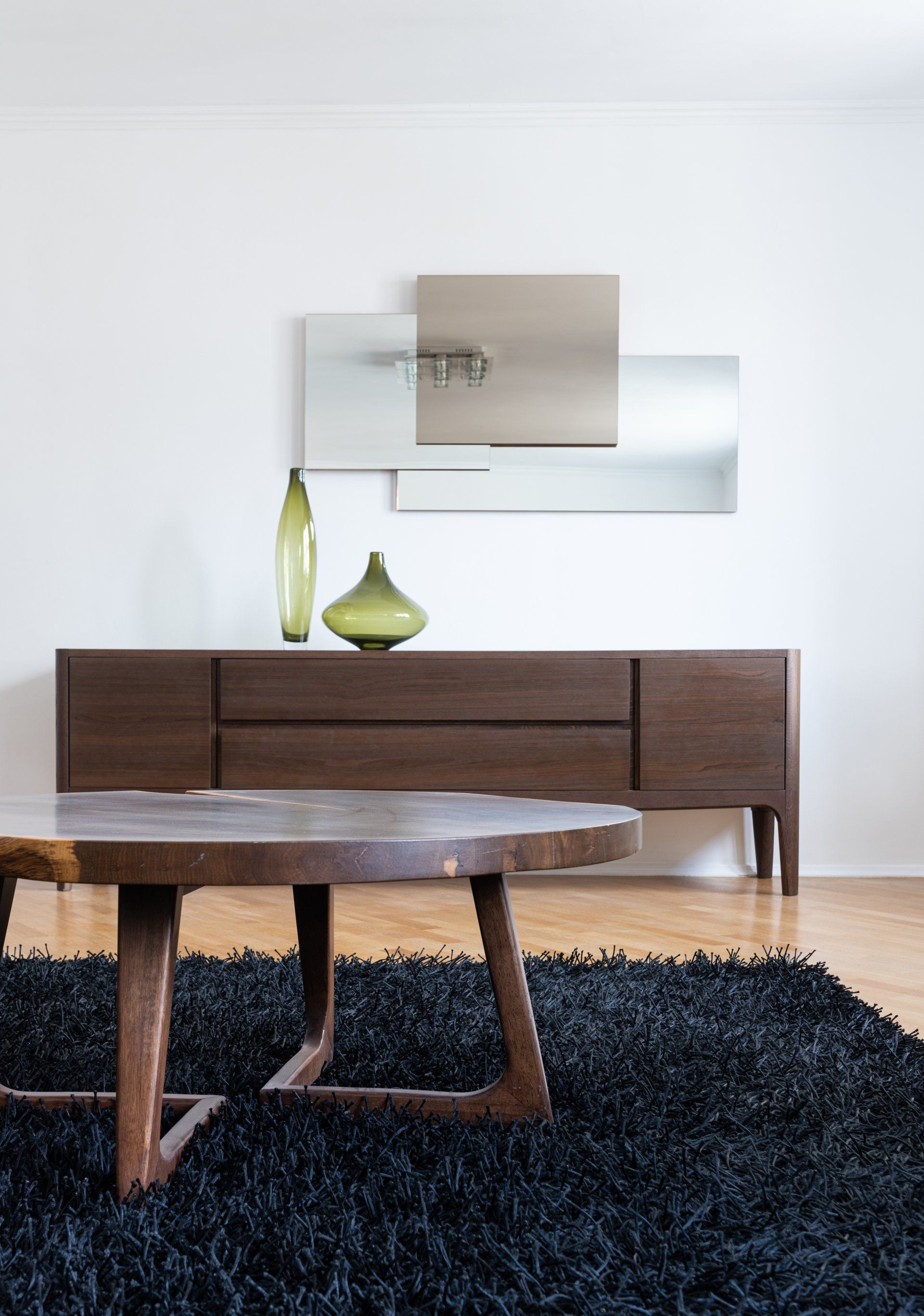 Modern living room with wooden sideboard, green vases, wall art, and round coffee table on a dark rug