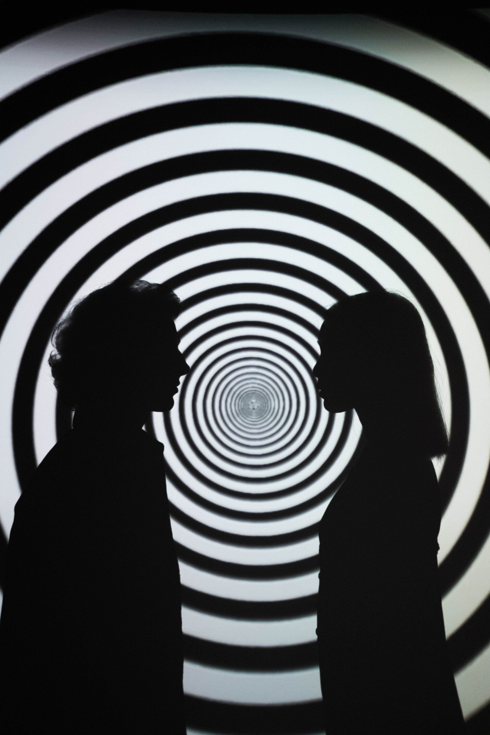 Silhouetted figures face each other before a black and white spiral backdrop.