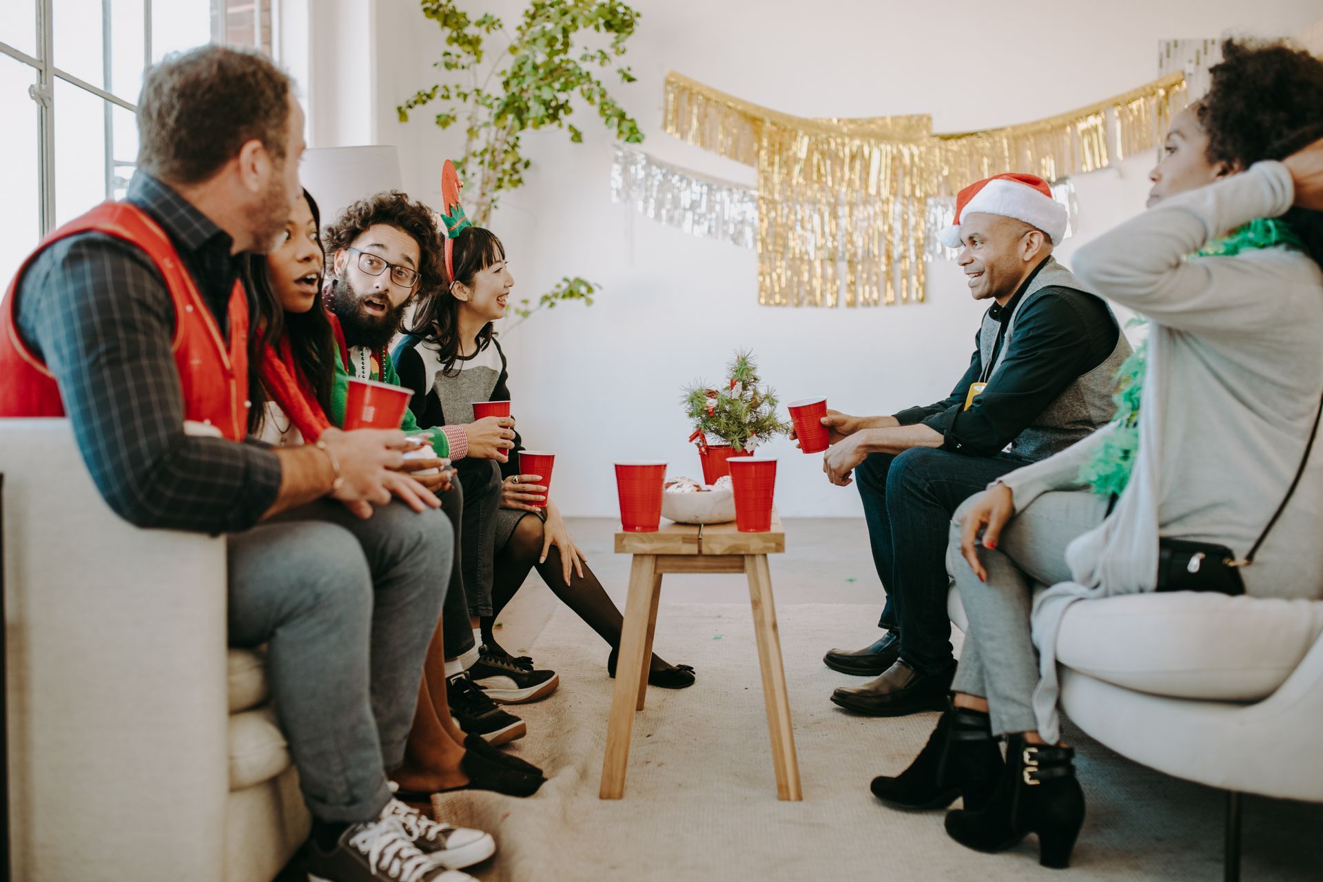 Group of people celebrating a holiday, sitting in a room, red cups, Santa hat, and festive decorations.