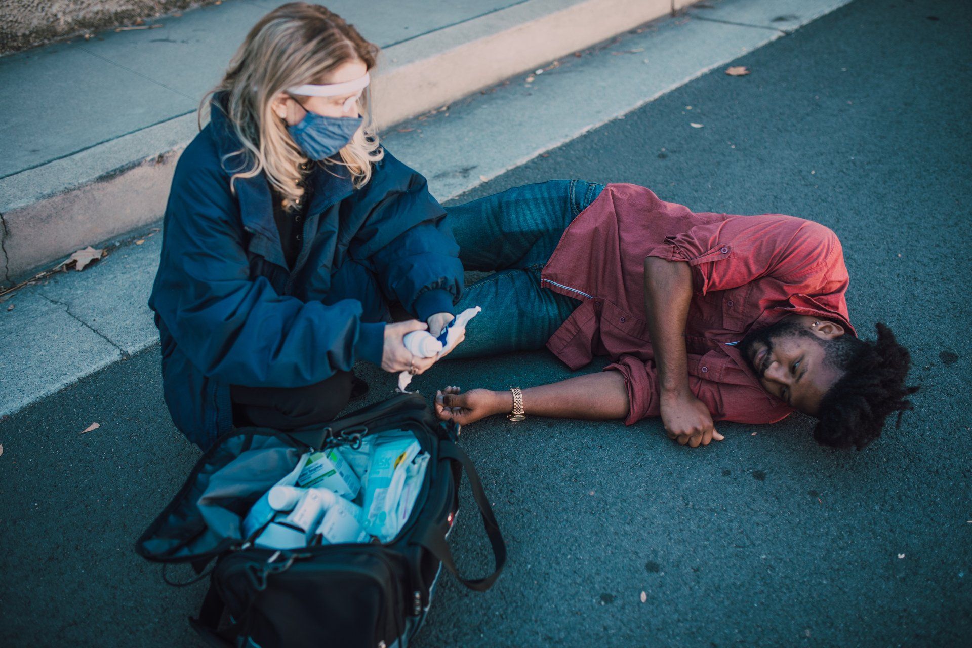 A woman in a mask tends to a man lying on the ground; she applies first aid. Both are on a paved road.