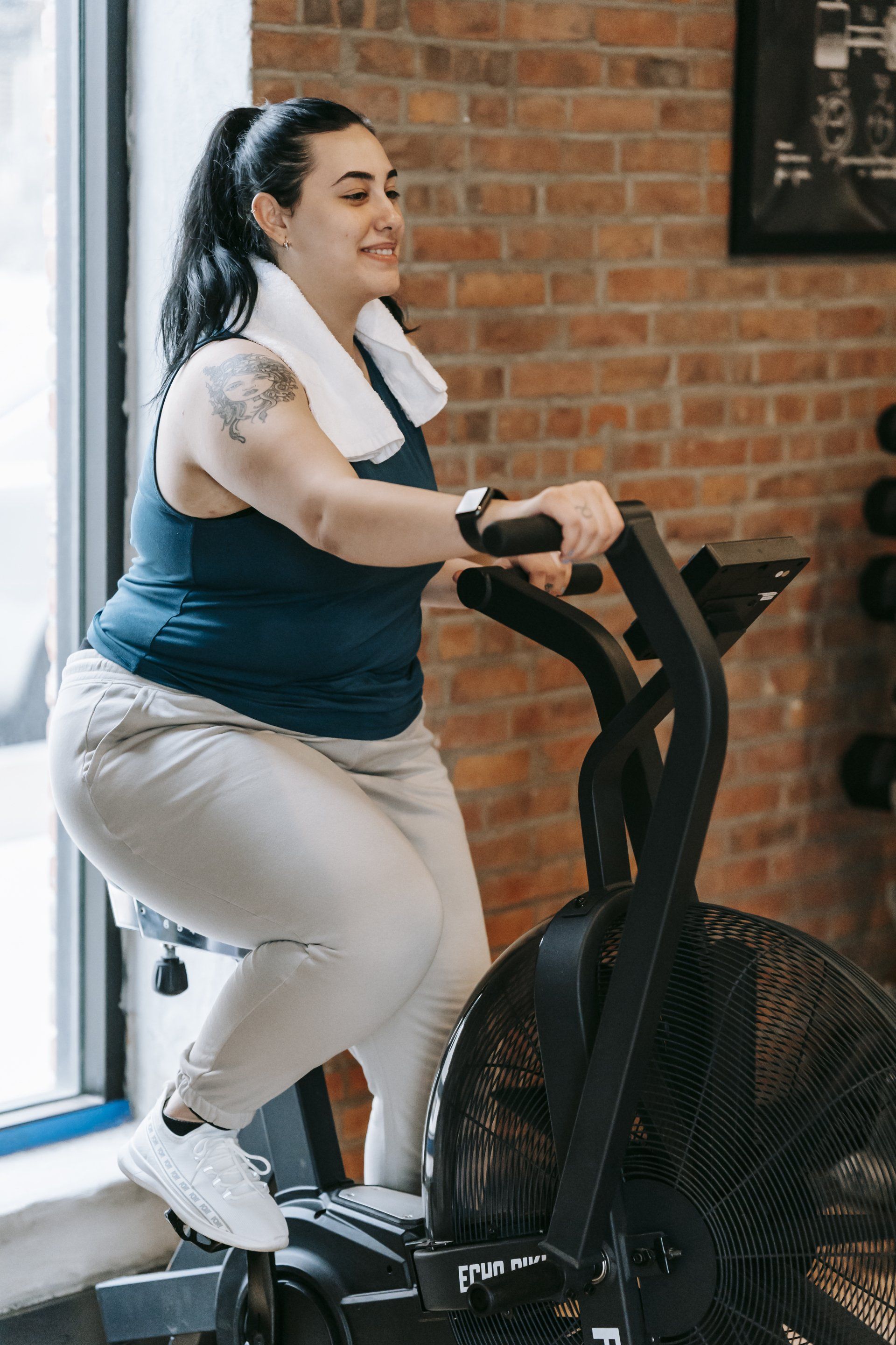 Person pedaling a black stationary bike in a brick-walled gym, wearing a navy top and light pants.
