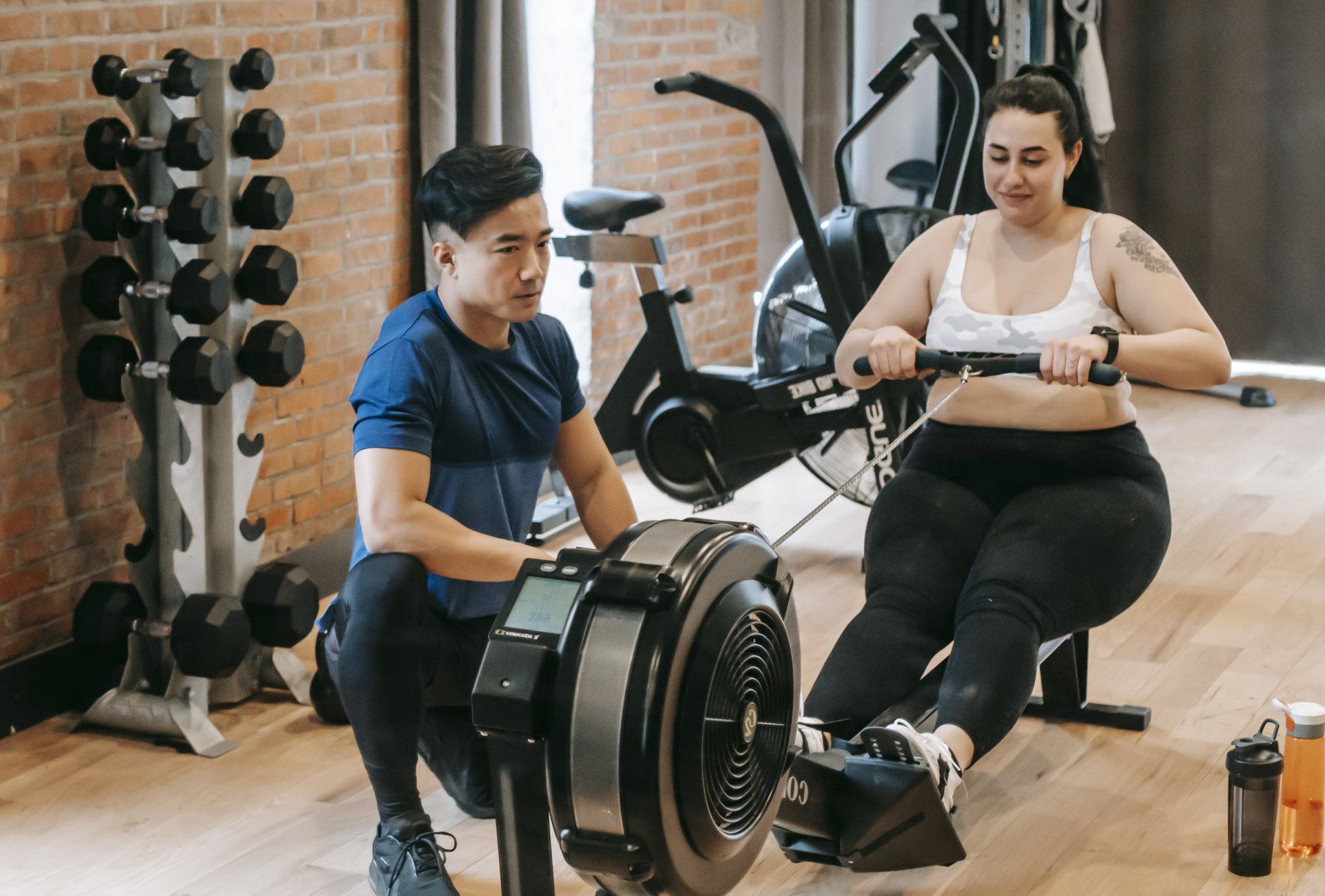 A woman rowing on ergometers in a gym, with dumbbells, exercise equipment, and a personal trainer or partner nearby.