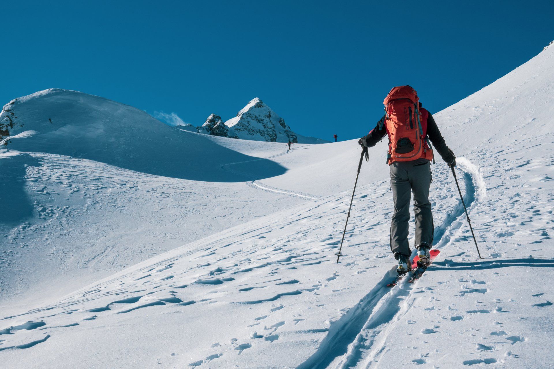 Skifahrer mit rotem Rucksack wandert unter strahlend blauem Himmel über einen schneebedeckten Berghang.