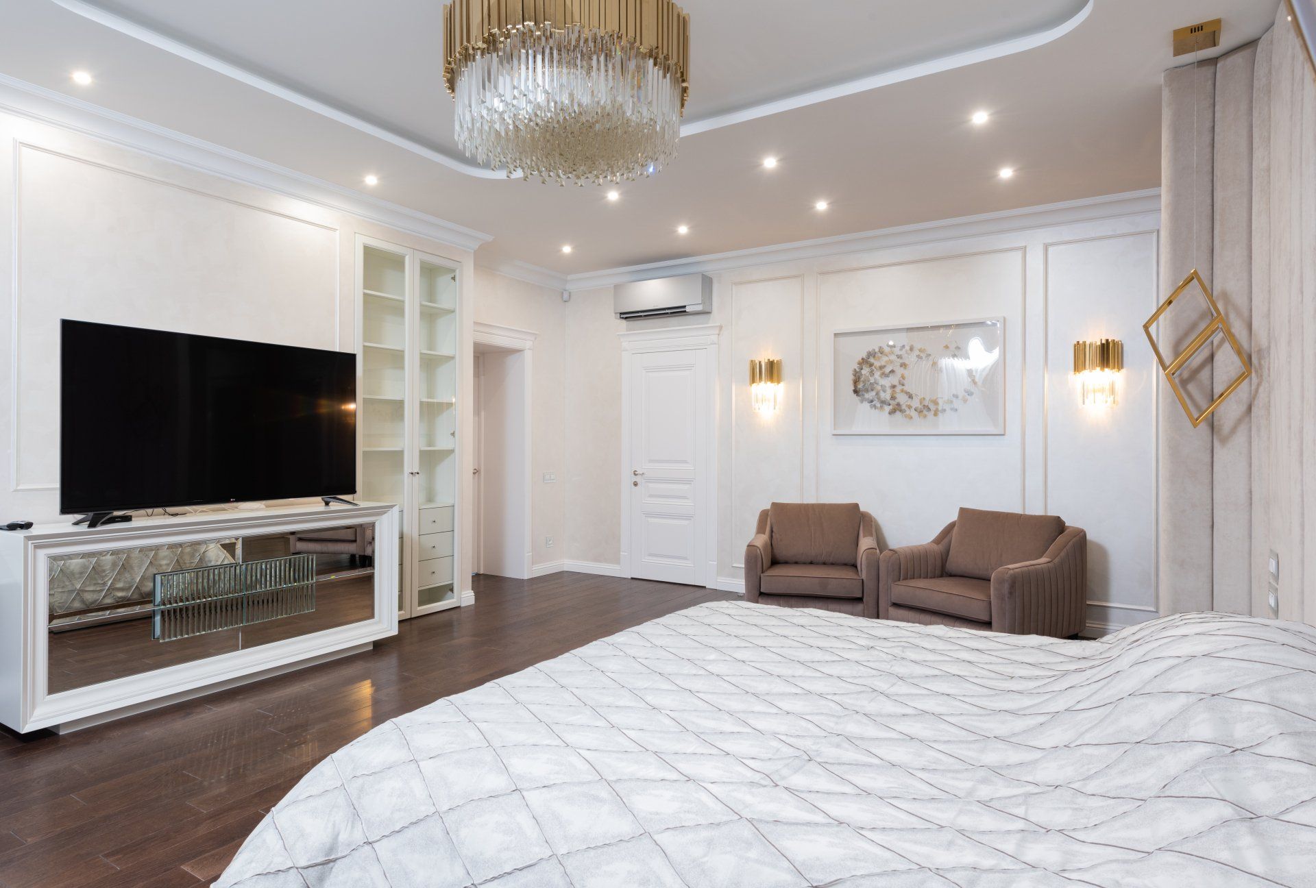 Elegant bedroom with white walls, chandelier, large TV, brown chairs, and a tufted bed spread