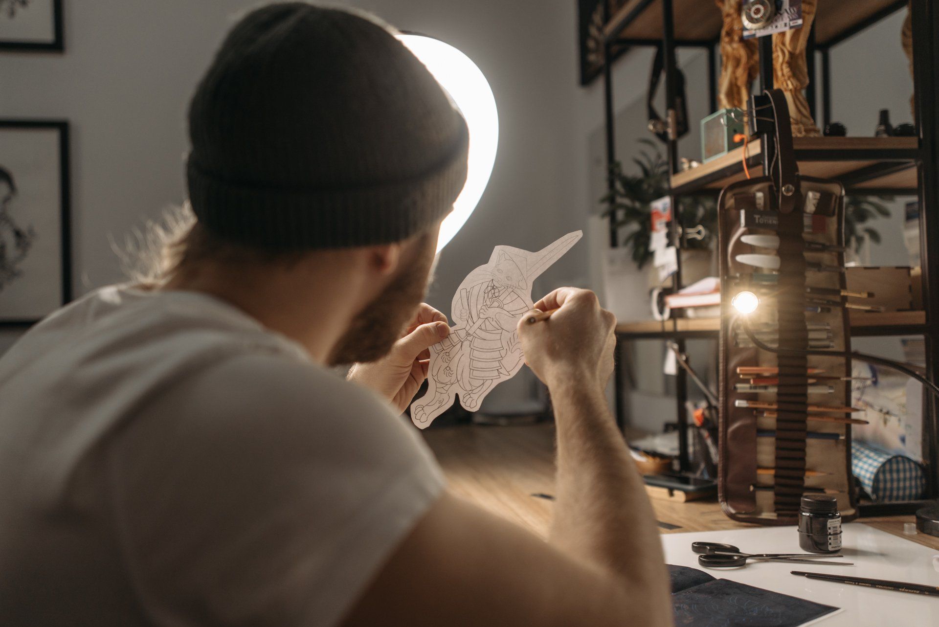Een persoon met een muts zit aan een bureau, verlicht door een fel licht, en bestudeert een gedetailleerde tekening op een uitgeknipte papieren sjabloon.