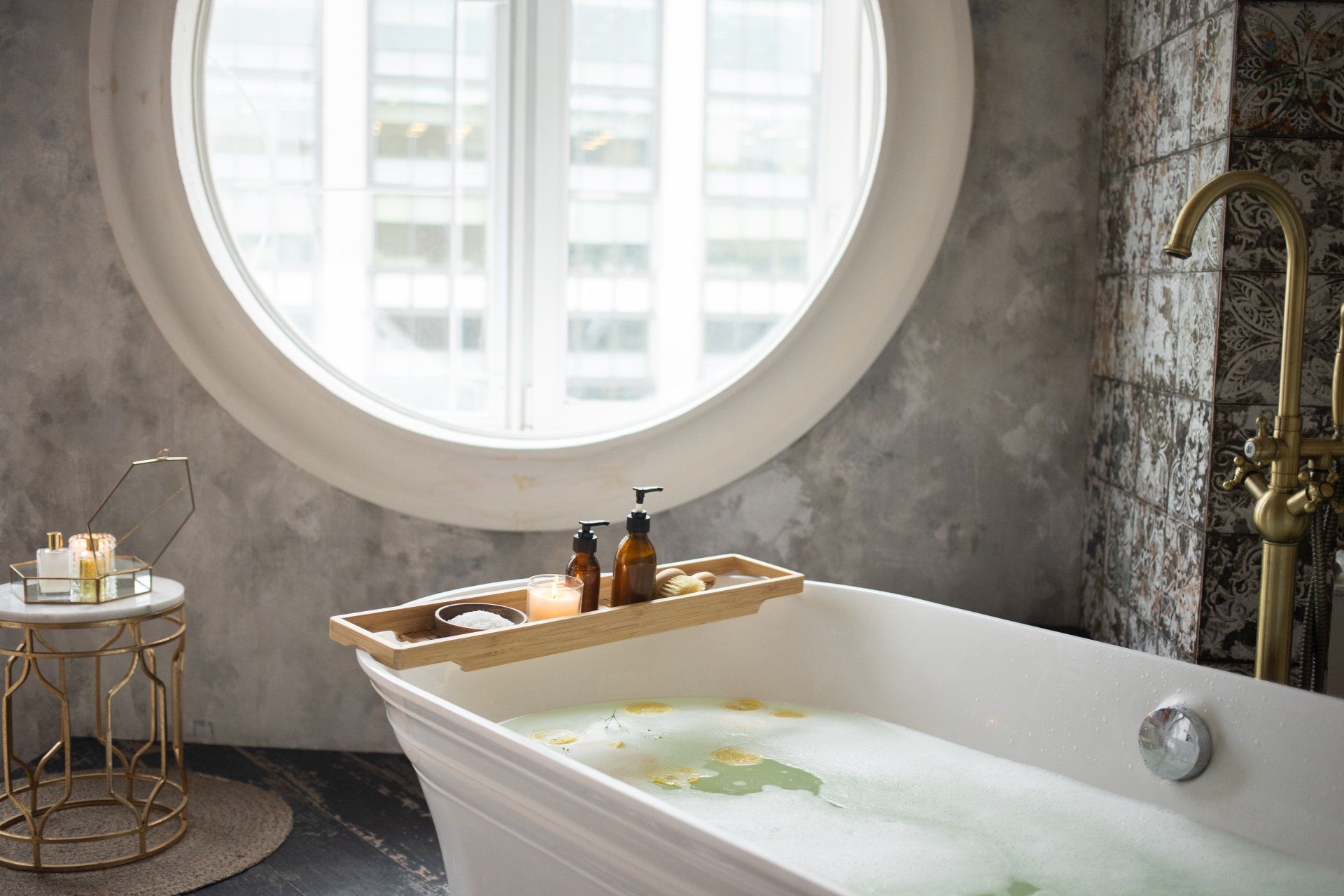 Freestanding bathtub beside a round window with stone wall and brass fixtures