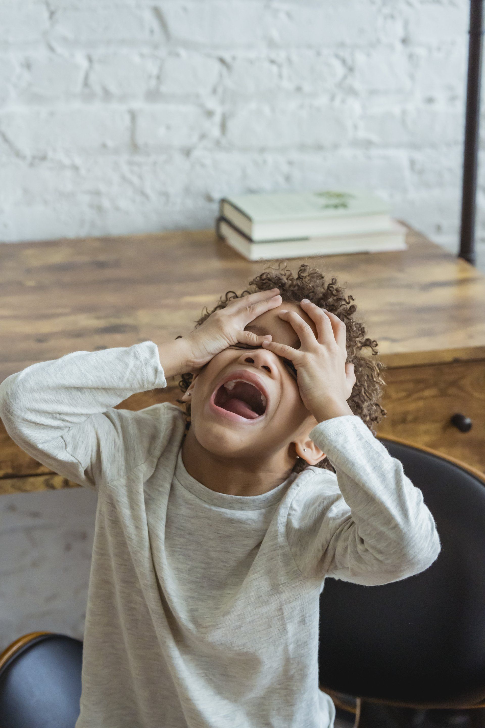 Child with curly hair covers eyes and cries, sitting at a desk with books.