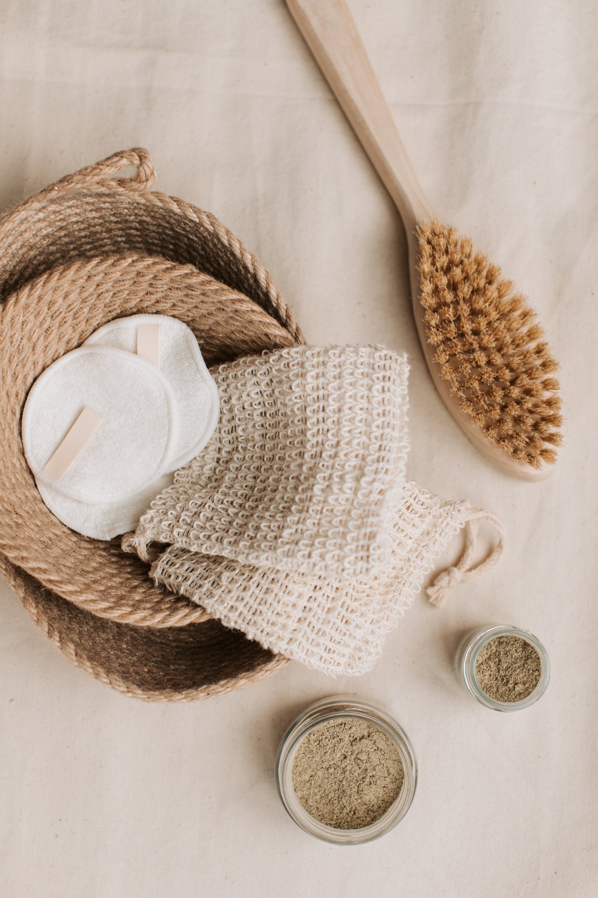 Woven hats, straw brush, knit tote, and small jars on a beige fabric background
