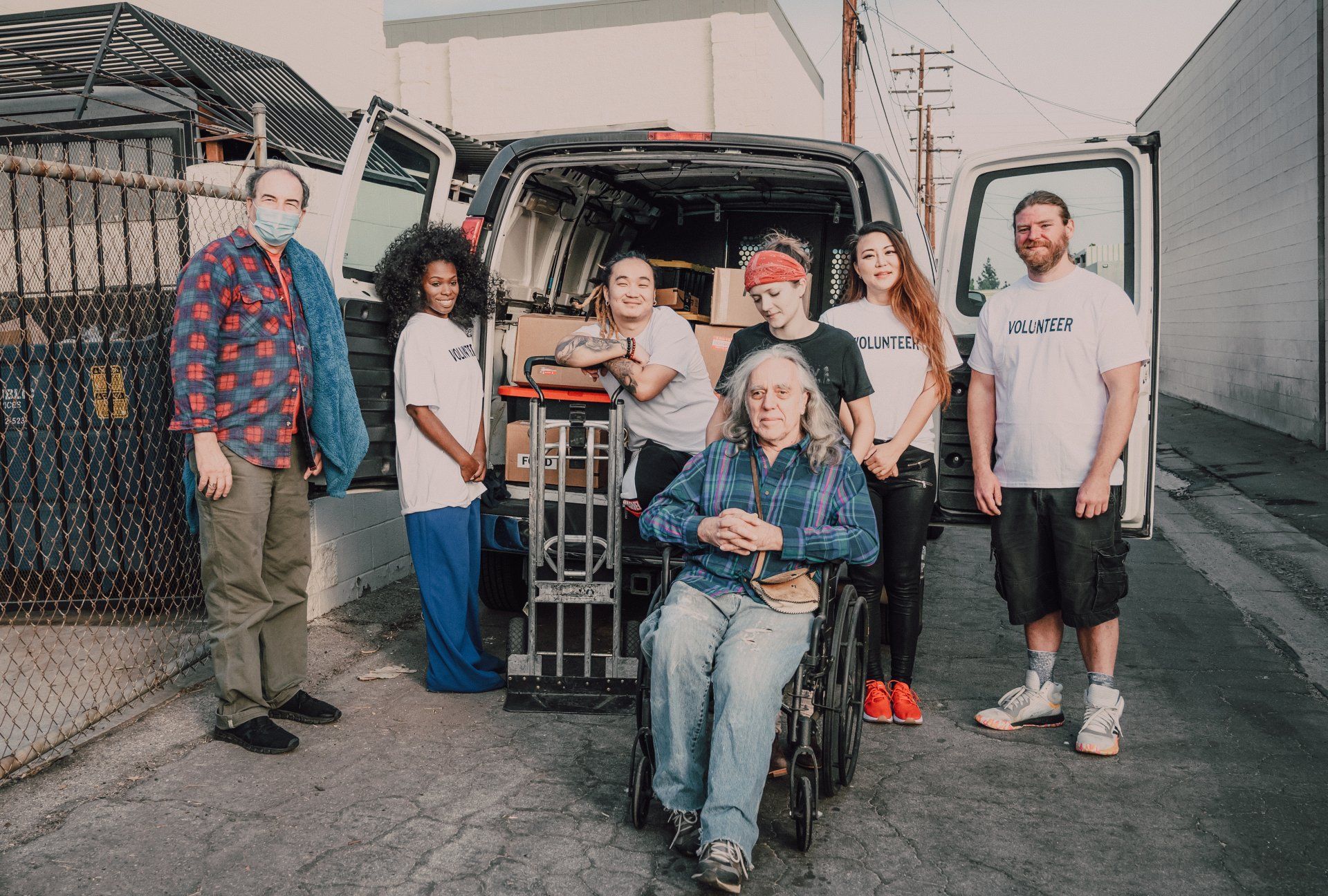 Group of people with a van, loading boxes. People smile. Outdoors setting, light gray pavement.