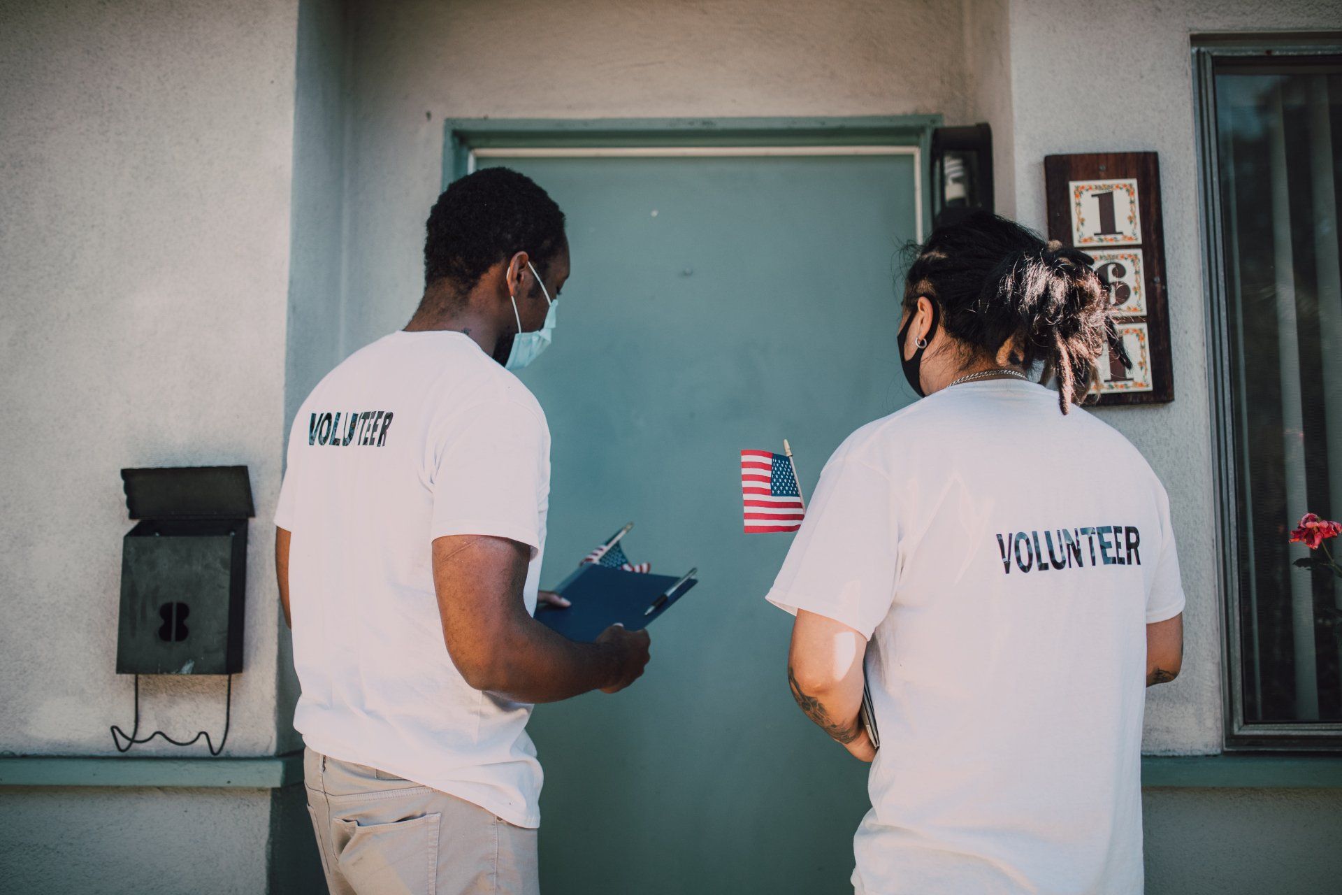 Two volunteers in white shirts speak outside a green door, one holding a clipboard.