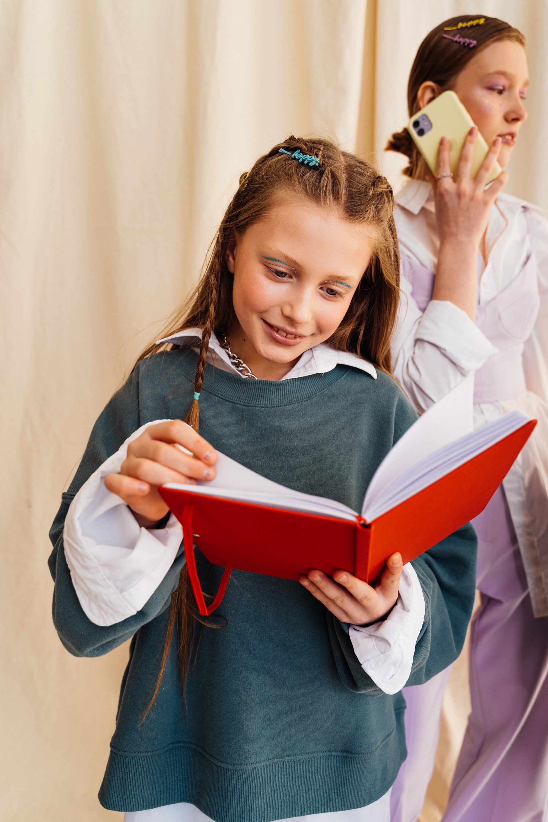 Child reading a red book while another person talks on a phone in the background
