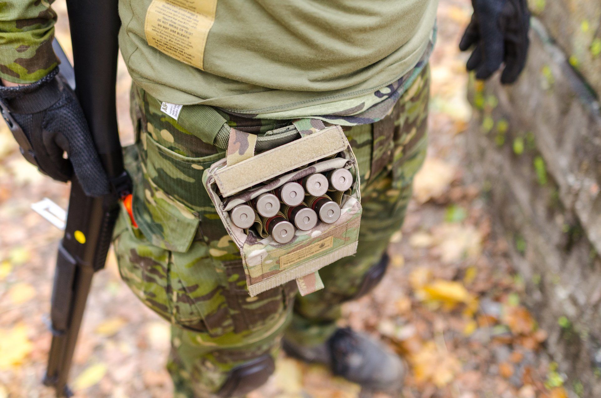 Camouflaged soldier holding a rifle, with a pouch of shotgun shells on a forest trail.