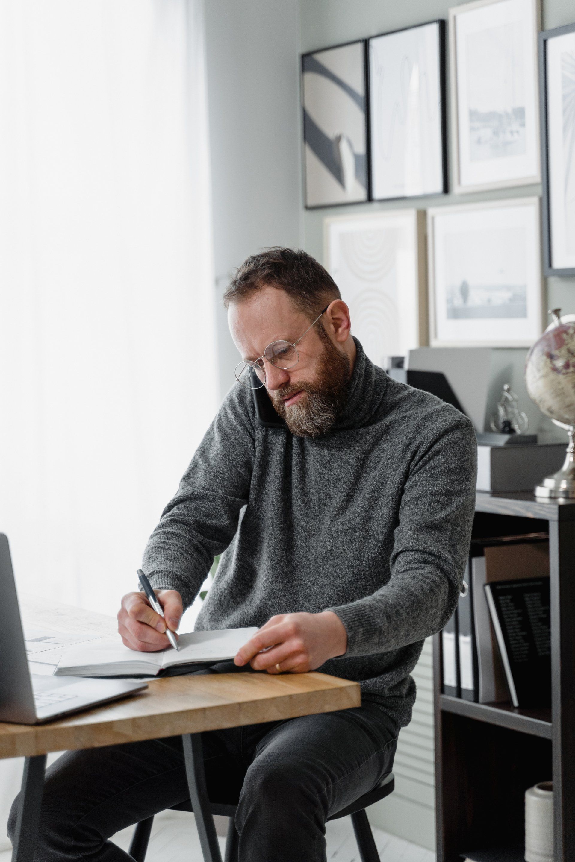 A man is sitting at a desk writing in a notebook while talking on a cell phone.