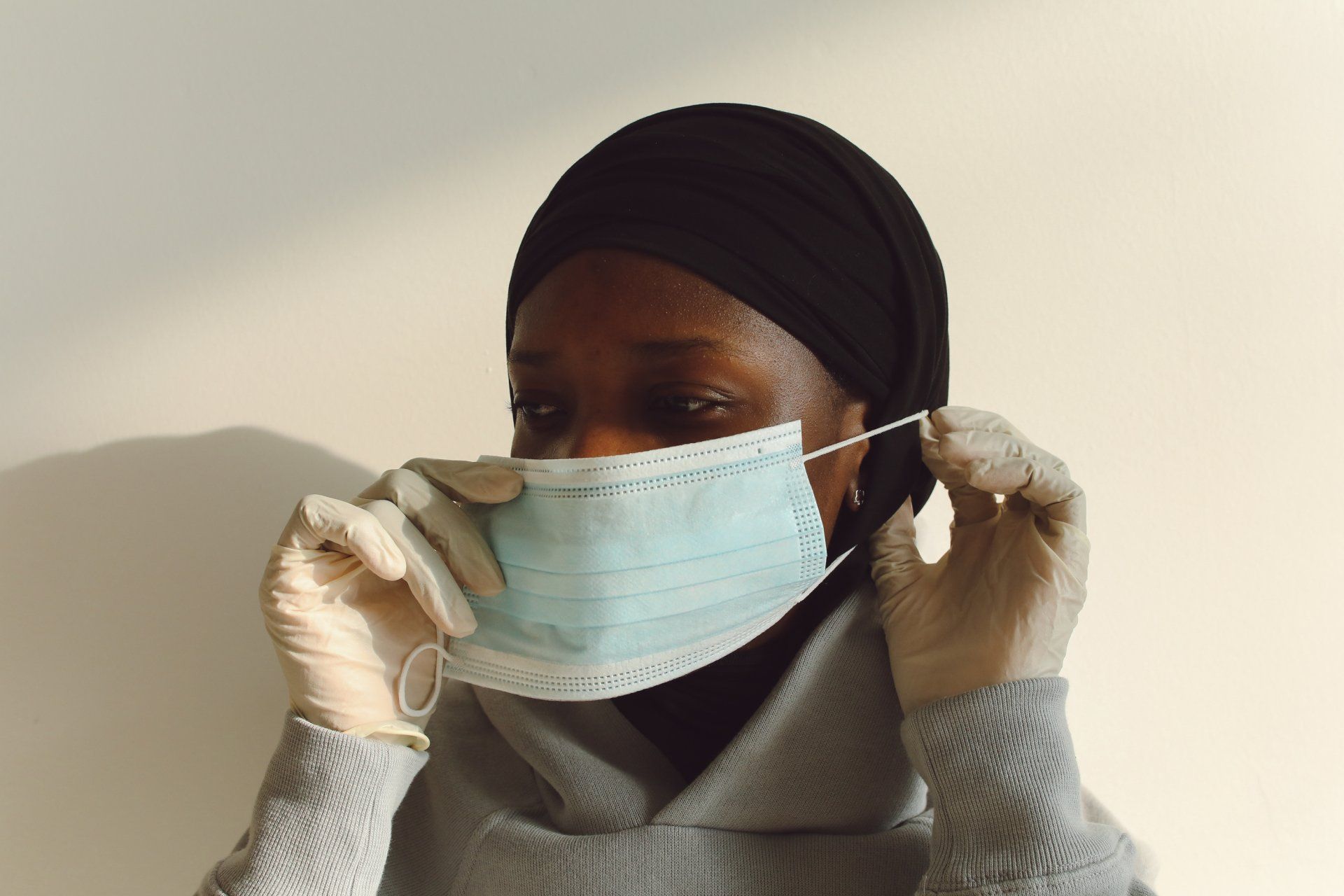 Person adjusting a light blue face mask while wearing white gloves and a black head covering
