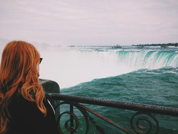 A person with reddish hair stands behind a metal railing, looking out at the cascading water of Niagara Falls.