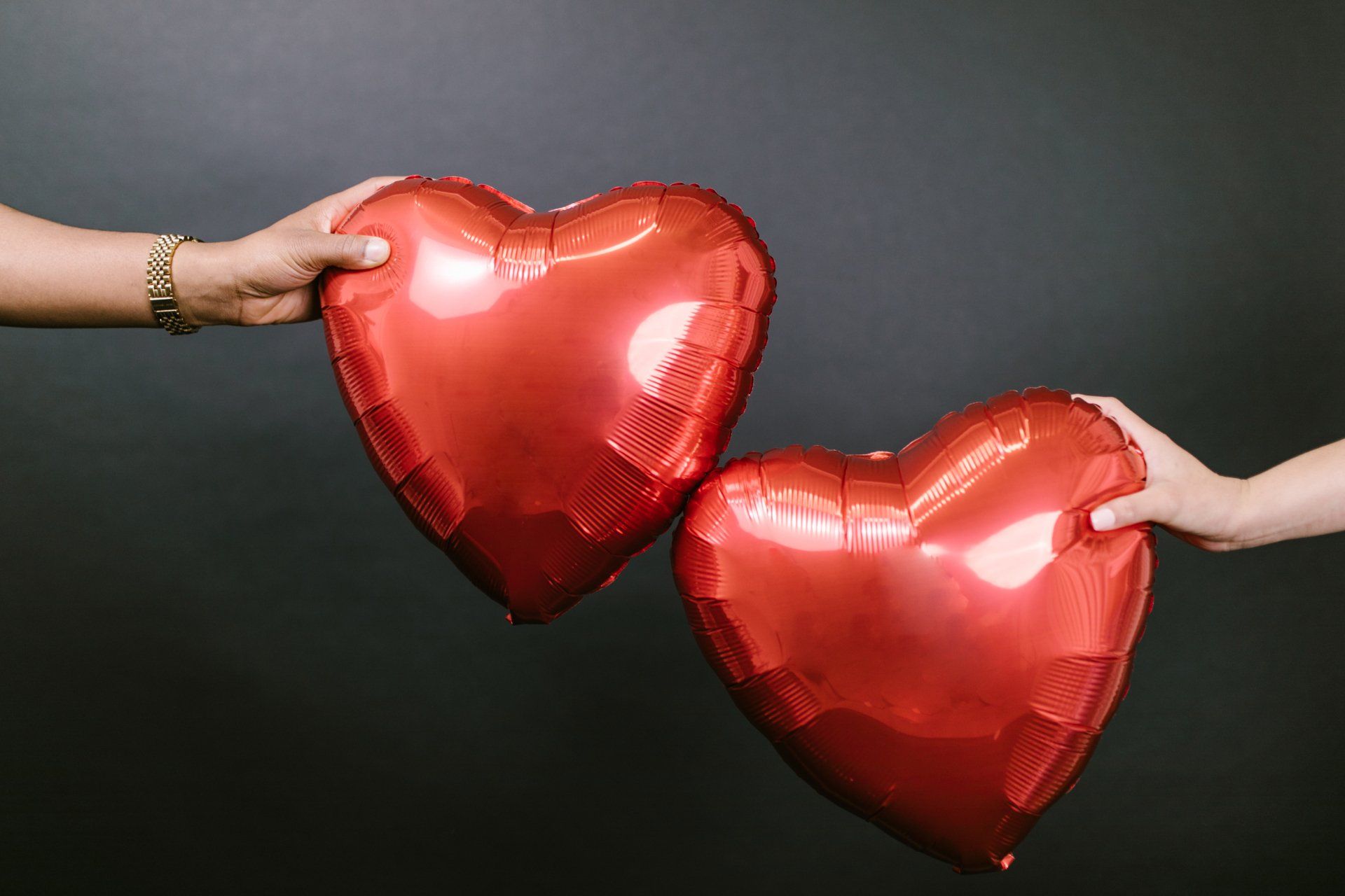 Two red heart-shaped balloons held by people's hands against a dark background.