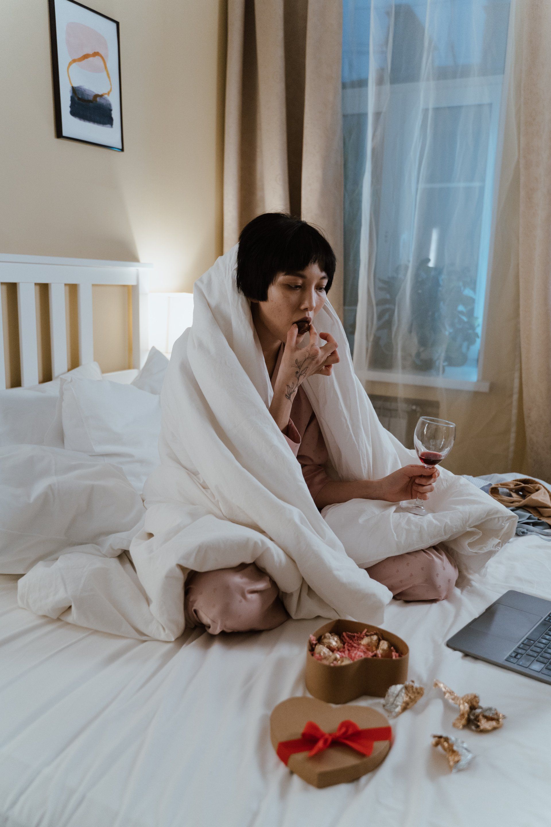 Woman in bed wrapped in blanket, sipping wine, with sweets, laptop.