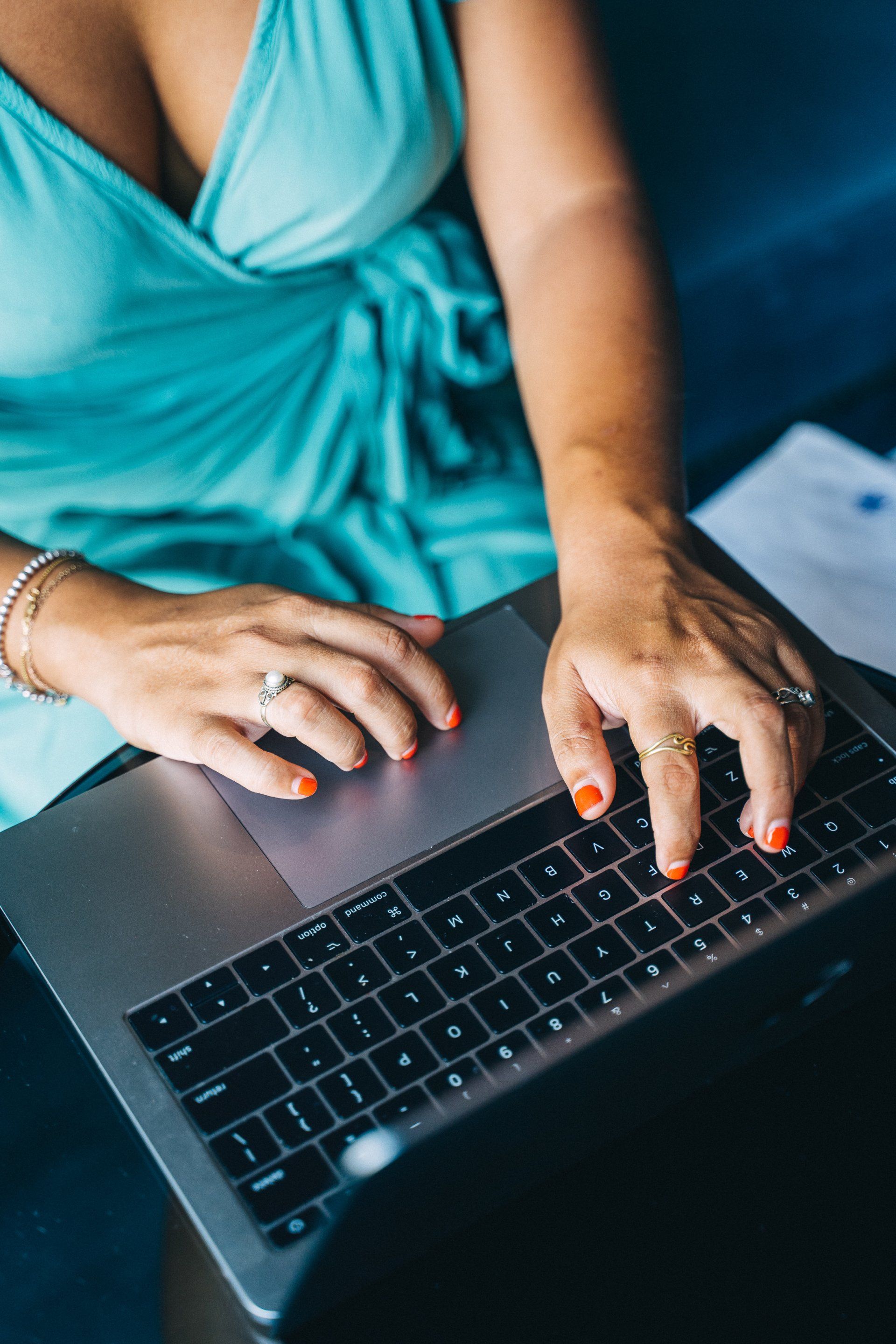 Woman typing on laptop; wearing teal wrap dress, orange nail polish, and rings.