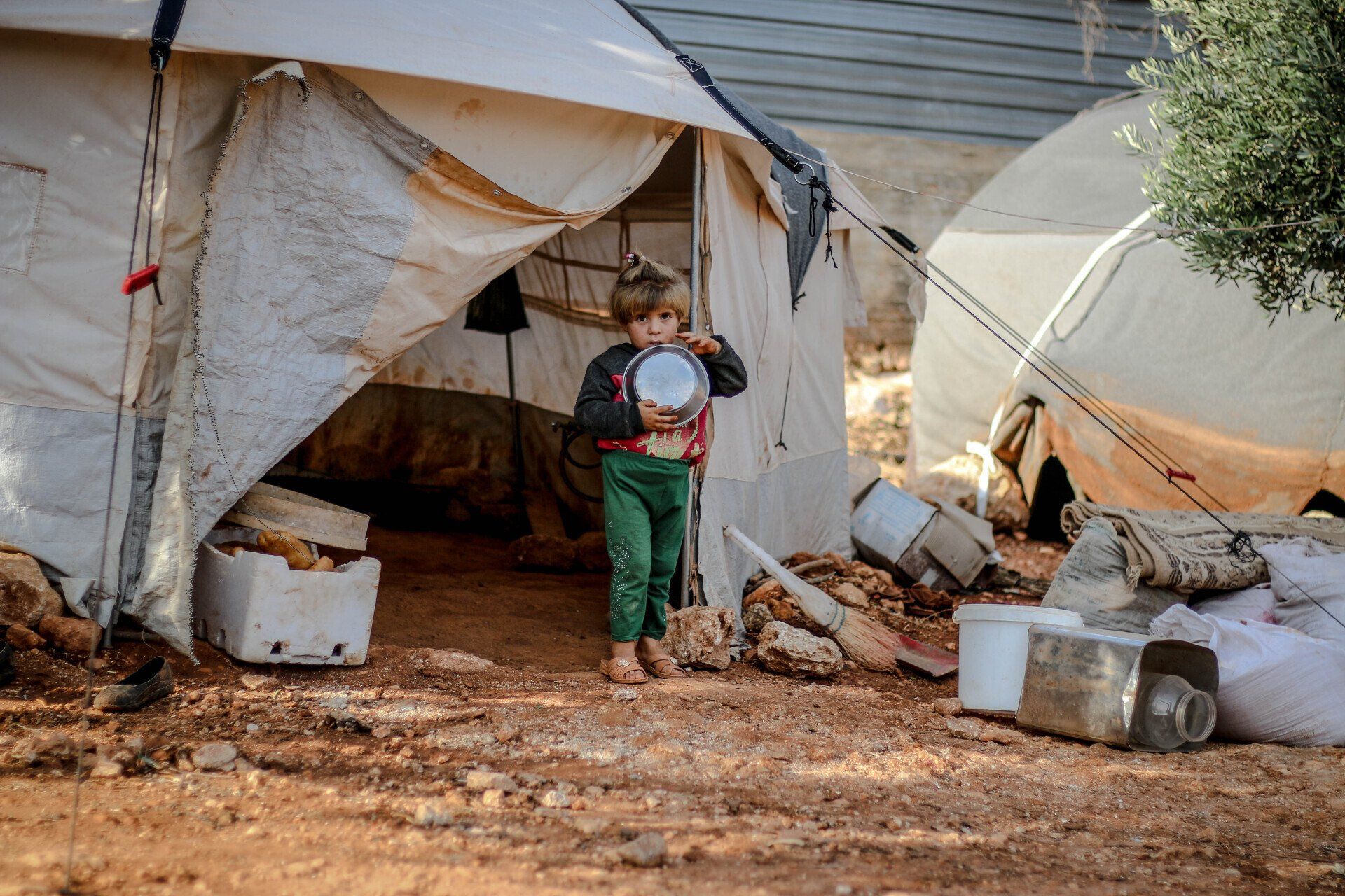 A child stands in front of a canvas tent, holding a small metal bowl in a sparse, outdoor setting. Chet's Roof charity and community service seattle
