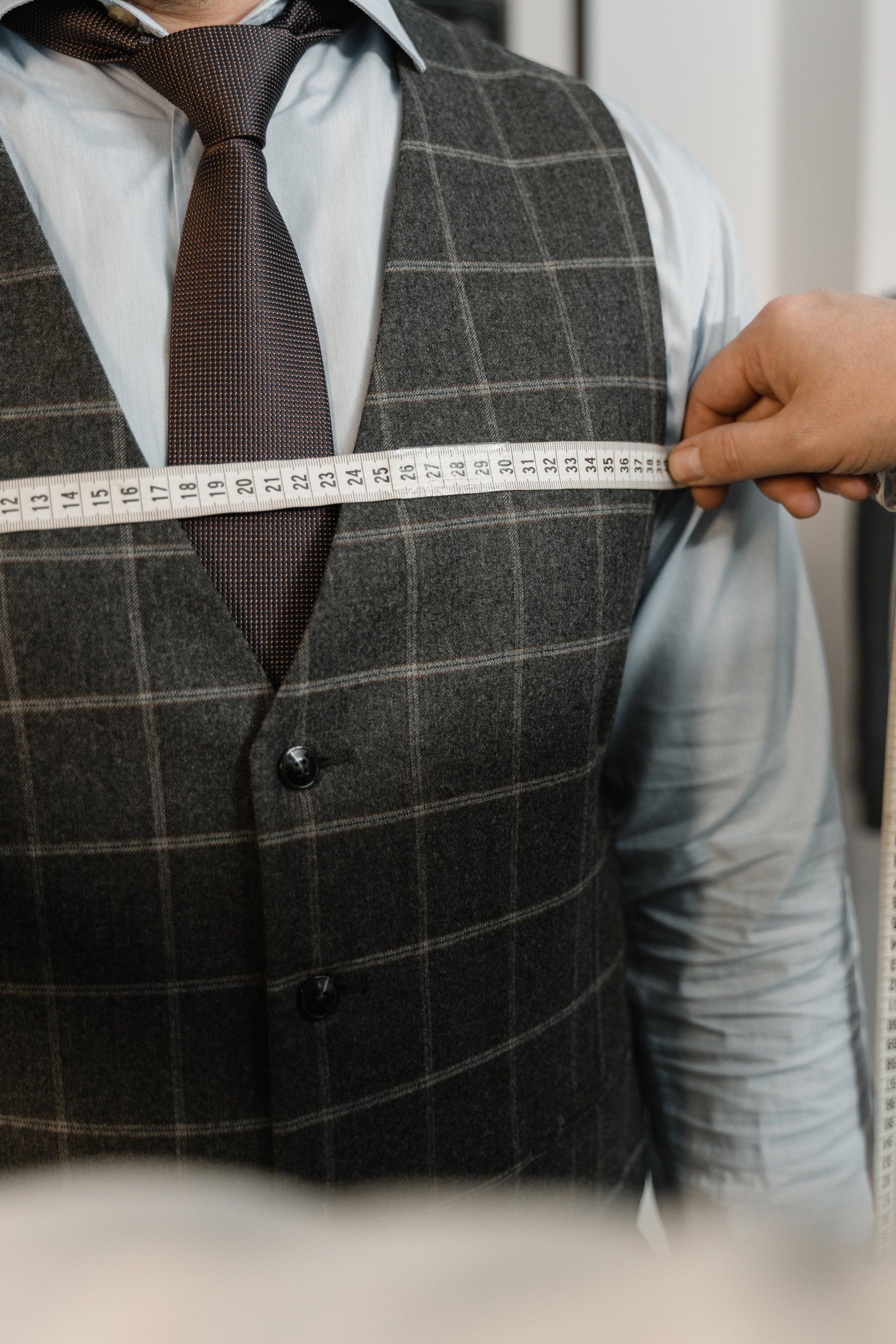 A person wearing a grey plaid vest and tie being measured for a suit with a white fabric measuring tape.