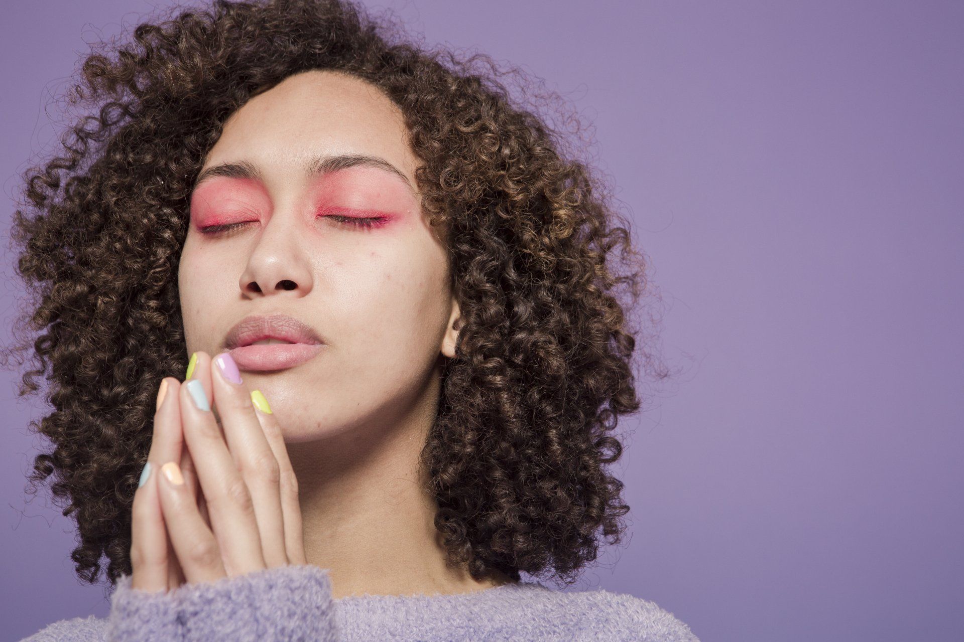 A woman with curly hair and pink eye shadow is praying with her eyes closed.