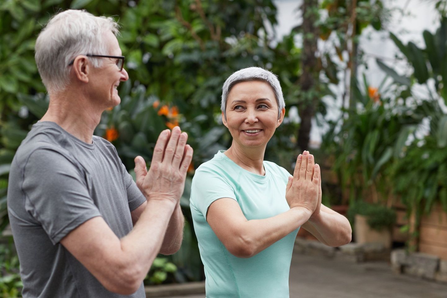 Older couple doing yoga, palms together, smiling outdoors in a garden.