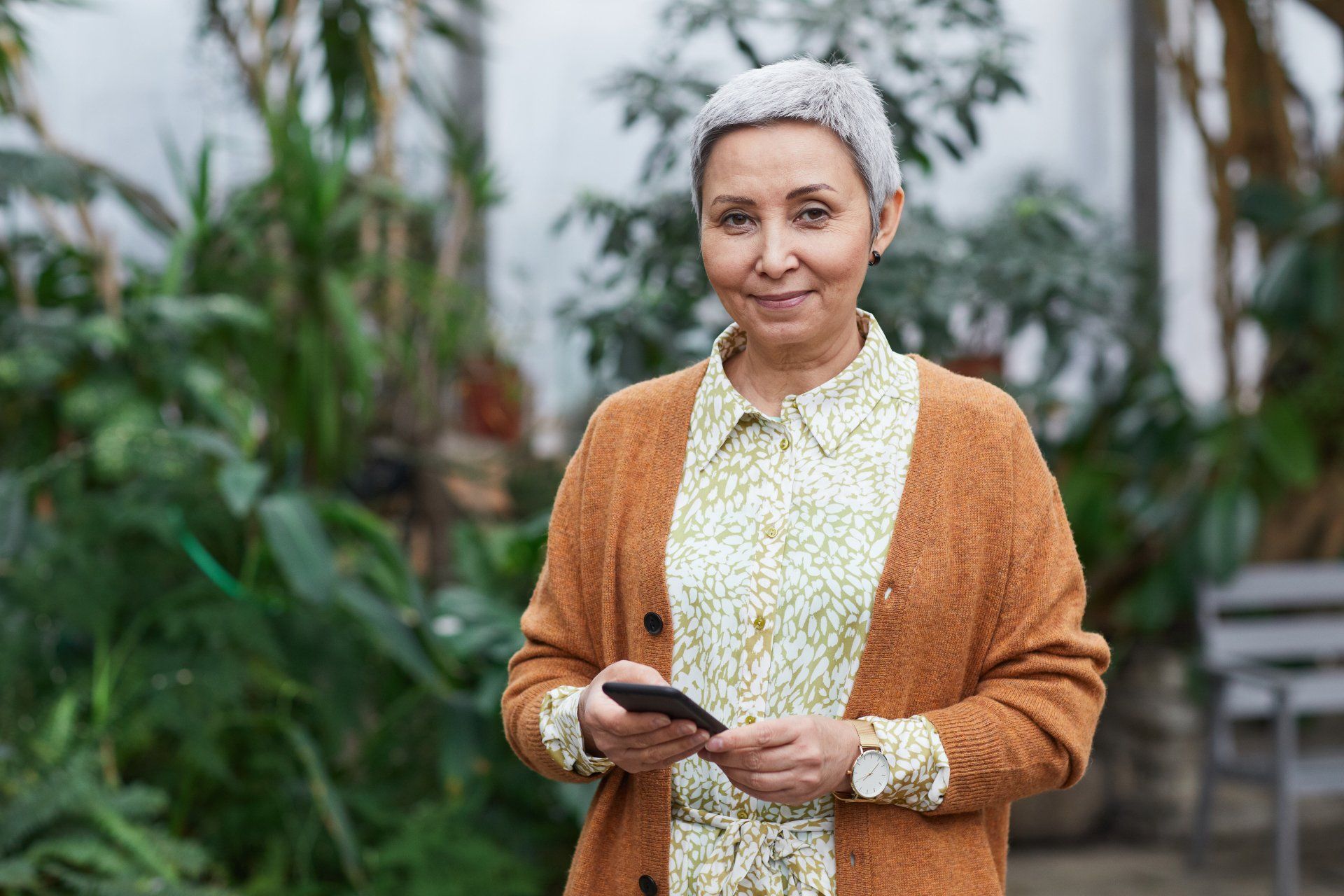 An elderly woman is standing in a garden holding a cell phone.