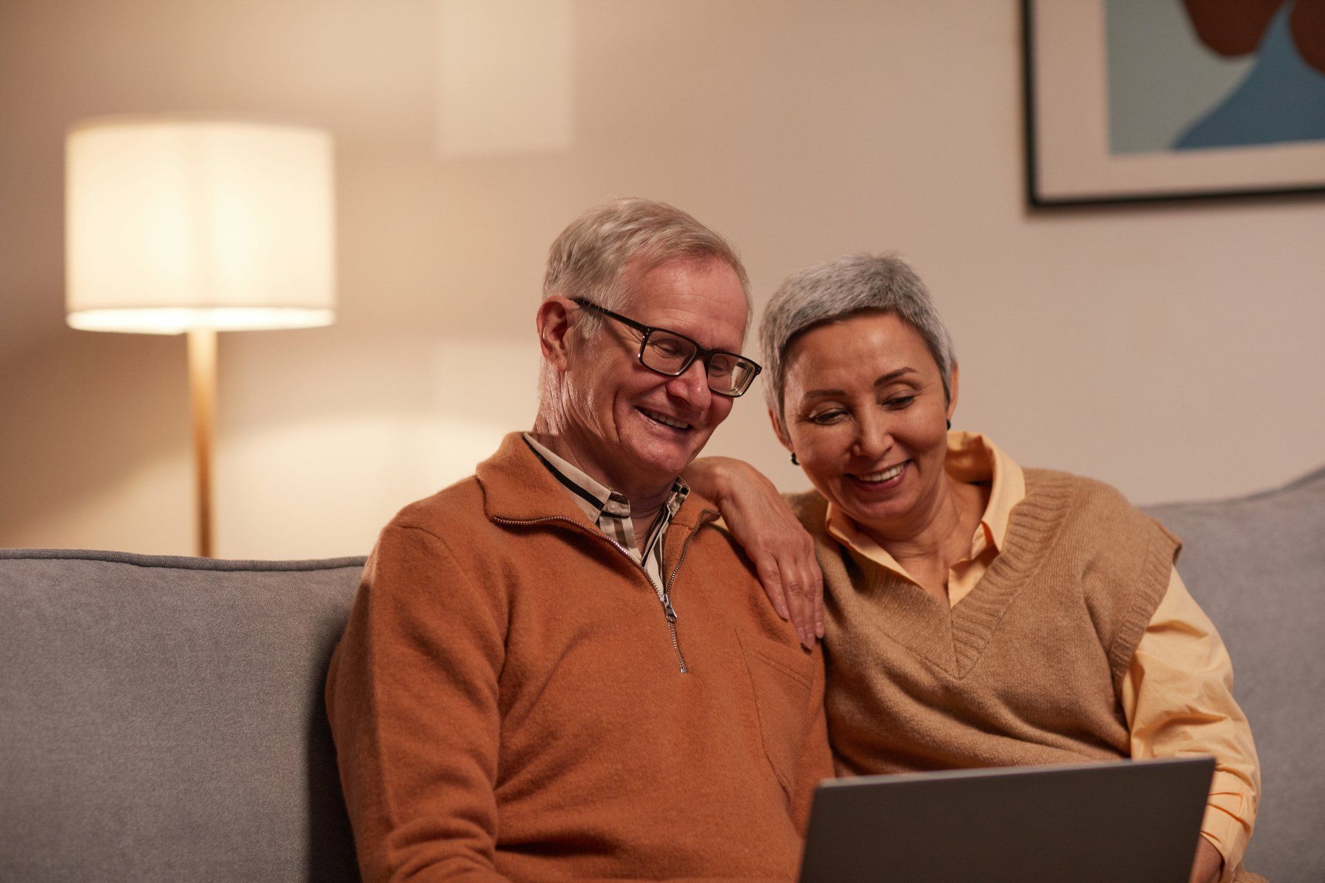 Elderly couple smiling, looking at a laptop together on a couch. Interior setting with a lamp and framed artwork.