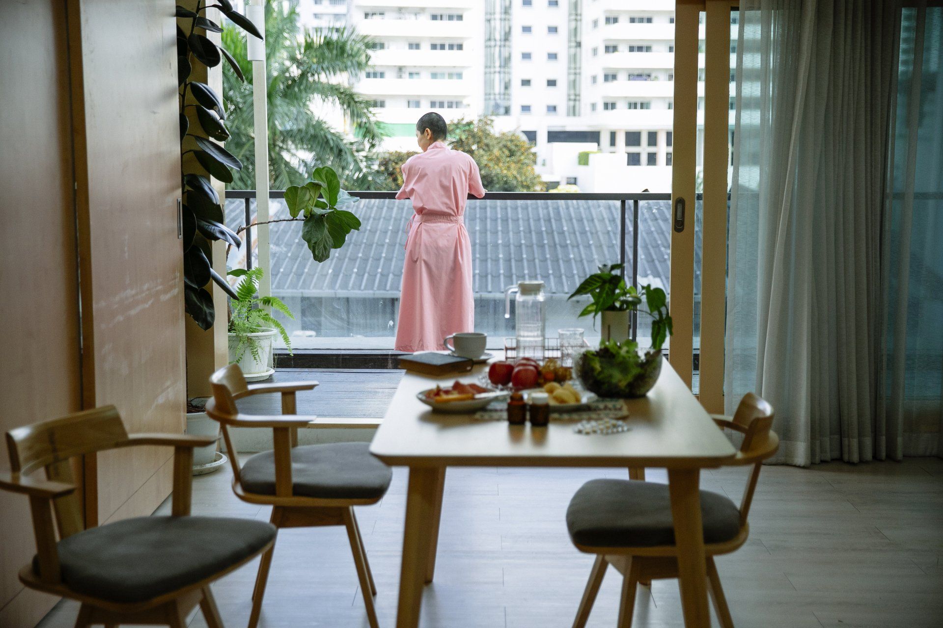 Person in pink dress on balcony, looking at city view, dining table set inside.