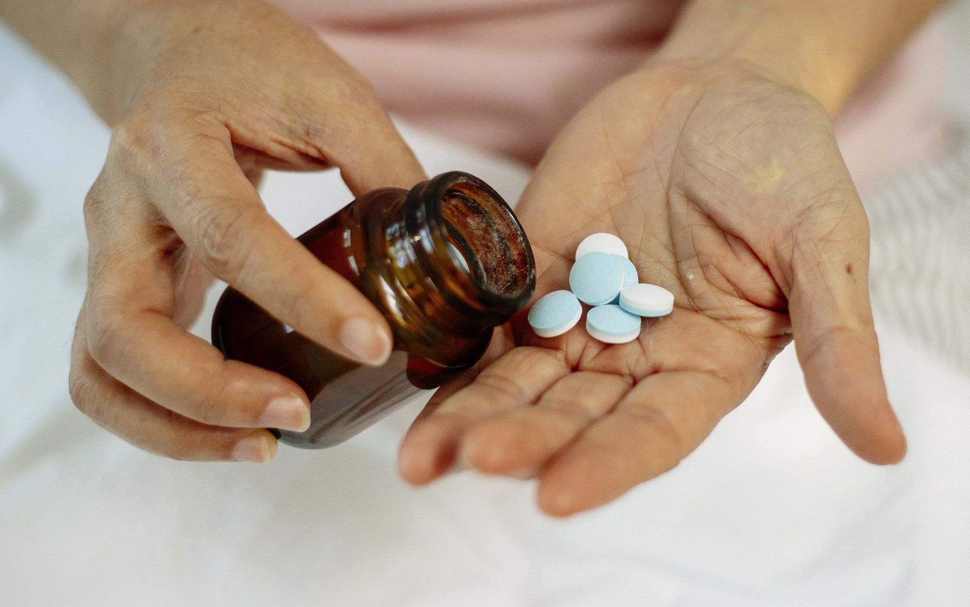 A person holds a small brown bottle and pours several light blue round pills into the palm of their hand.