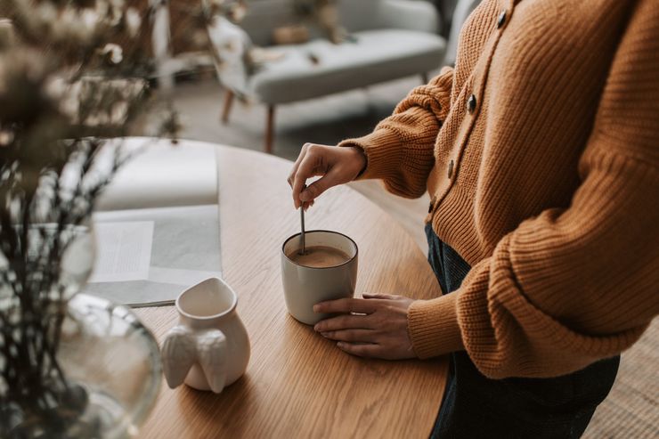 Person stirring coffee in a mug on a wooden table, wearing an orange sweater.