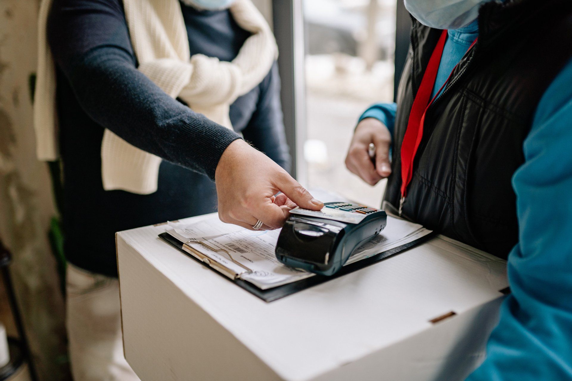 Person using a card to pay a delivery person at a doorstep. The card is tapped on a payment device held over a package.