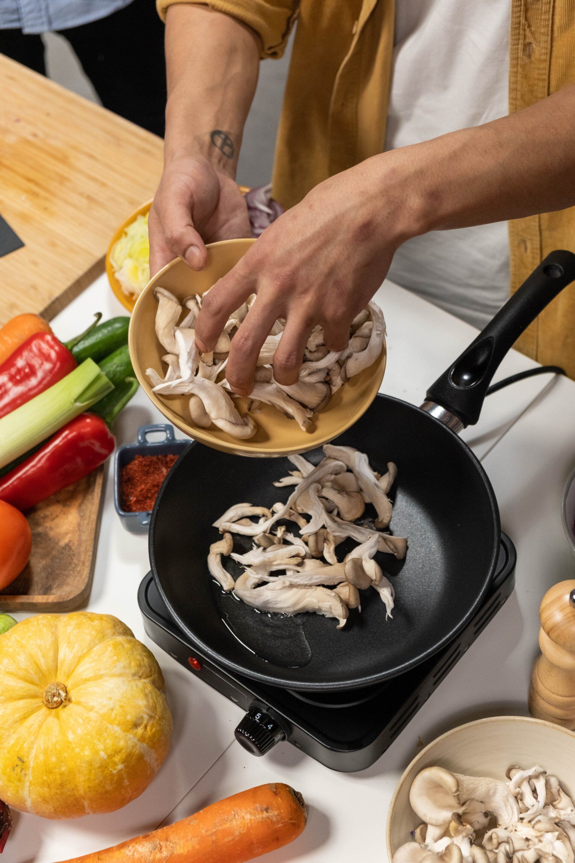 Person adding sliced mushrooms to a pan on a portable burner; vegetables and ingredients surround.