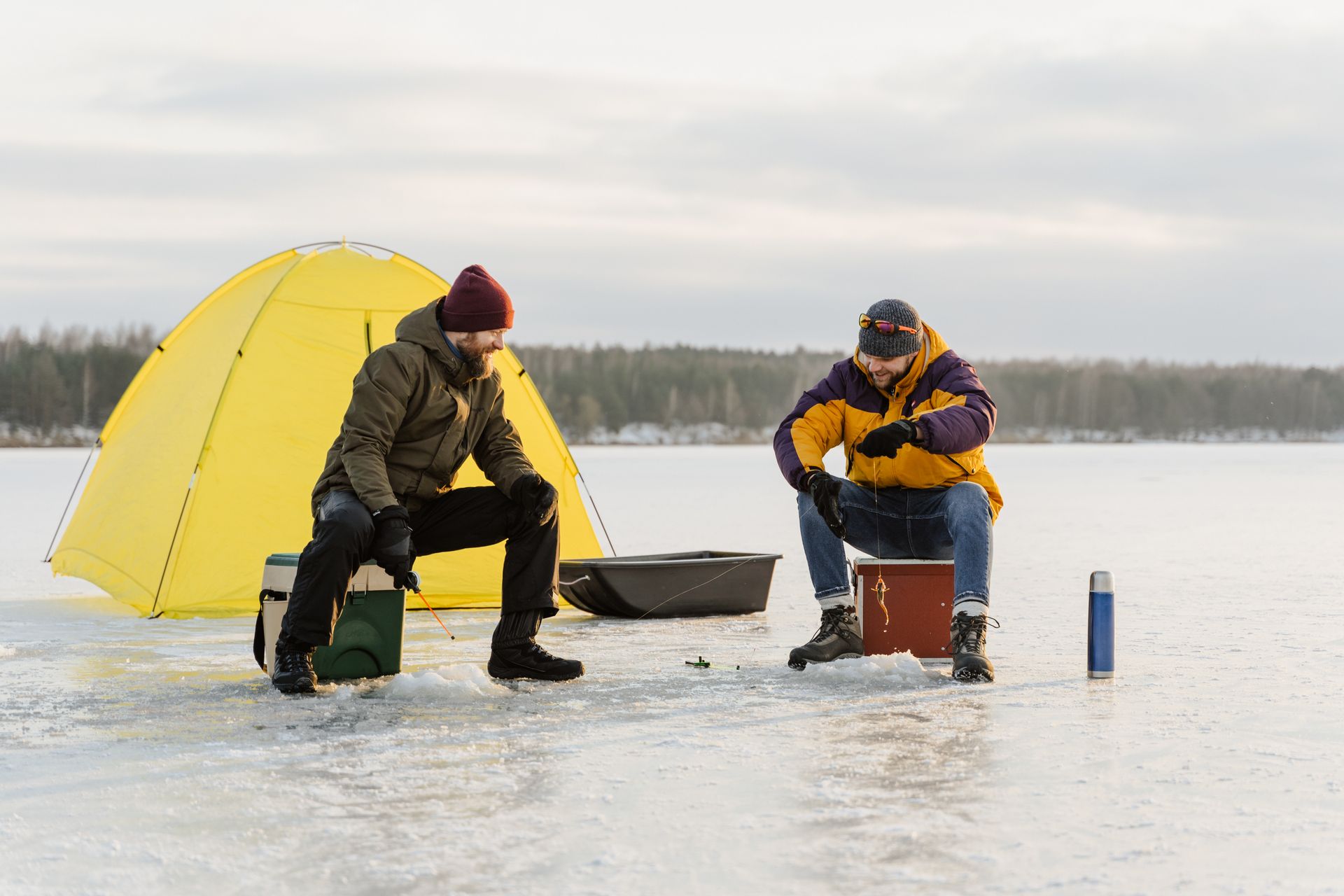Two men are ice fishing on a lake with a yellow tent in the background.
