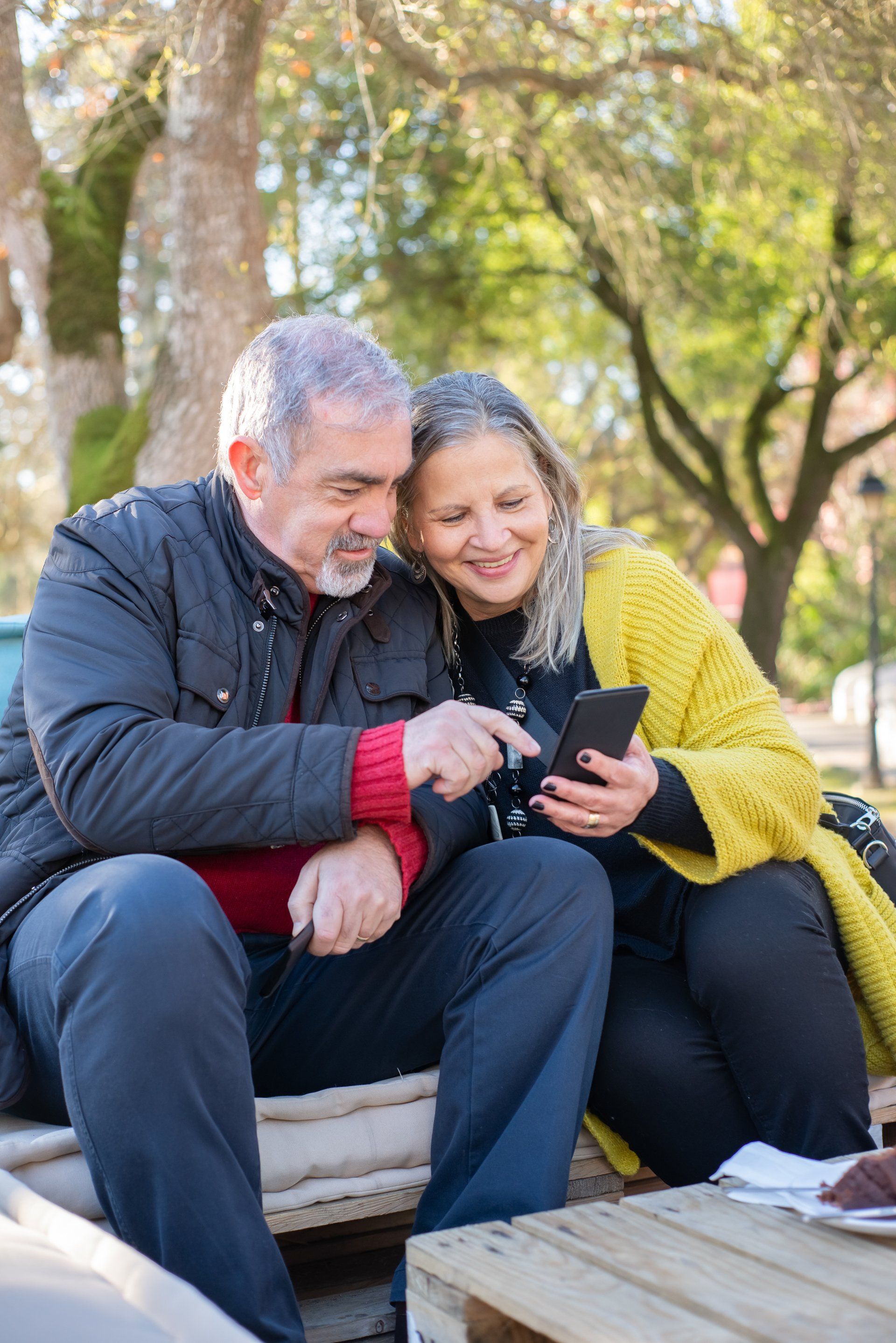 Couple looking at a smartphone, smiling. Outdoors, sitting on a bench.