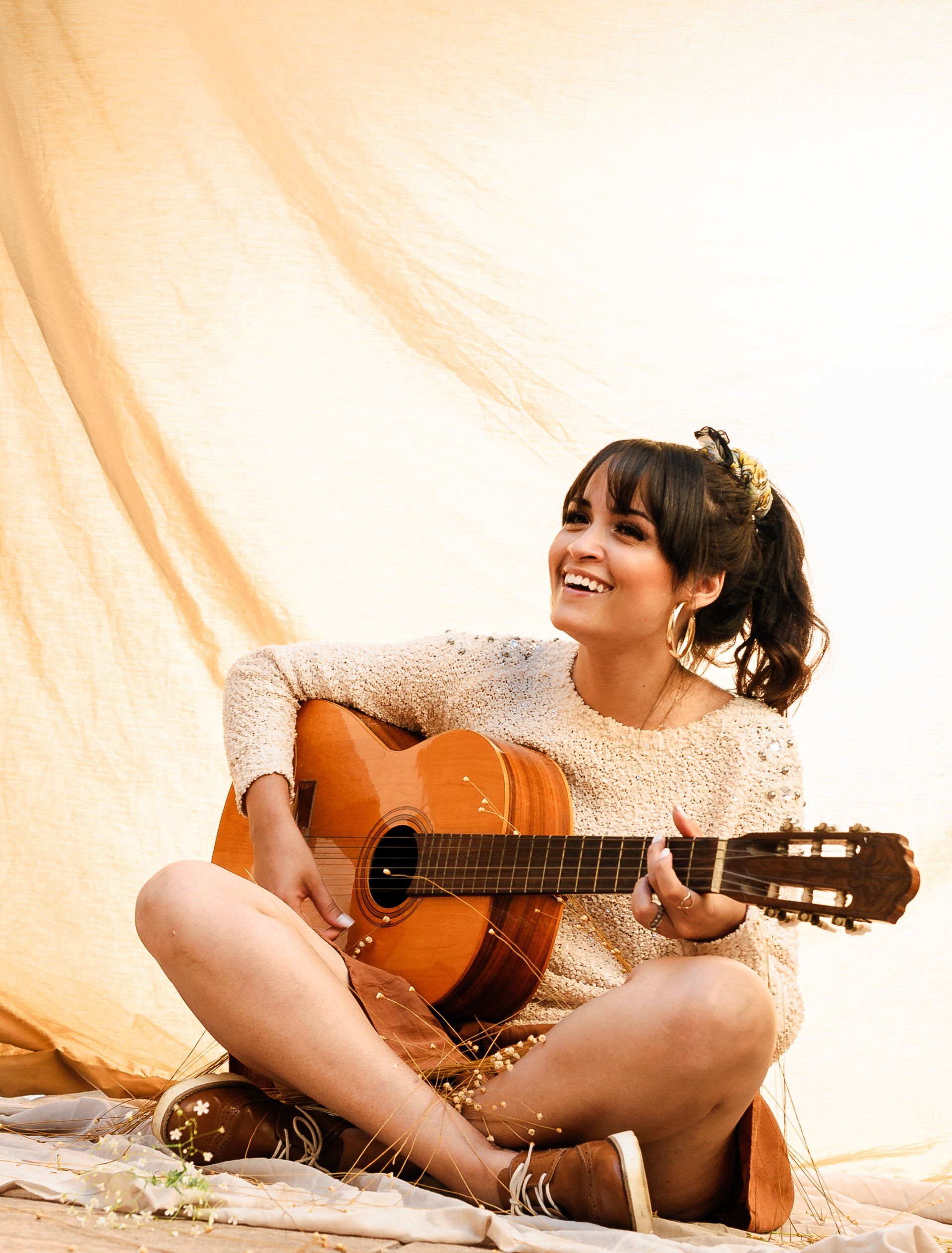 Woman sitting on a floor with an acoustic guitar, smiling in a warm, sunlit studio setting