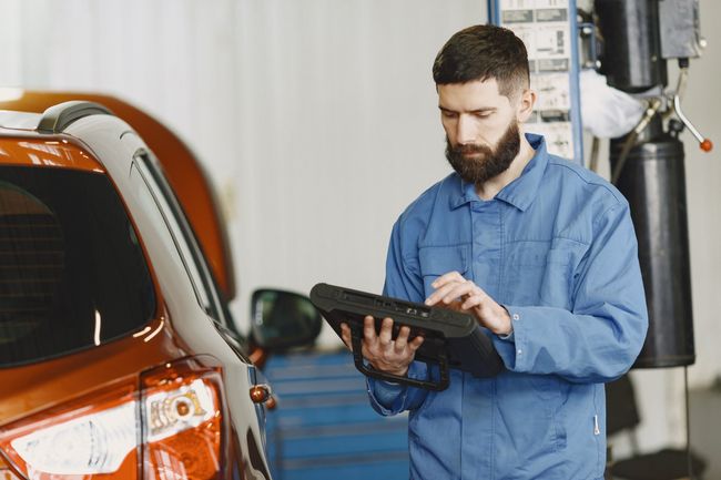 Mechanic in blue coveralls using a tablet beside an orange car in a garage
