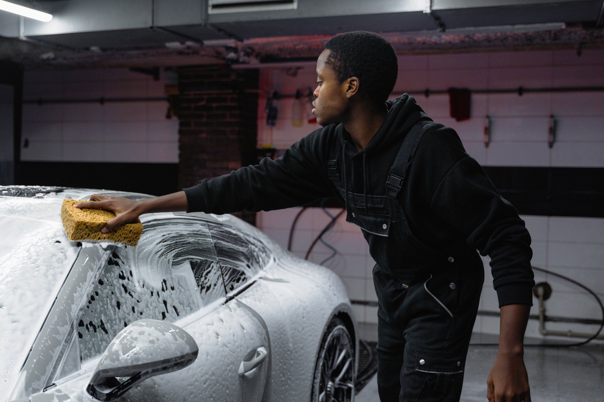 Person washing a silver sports car in a garage with a yellow sponge
