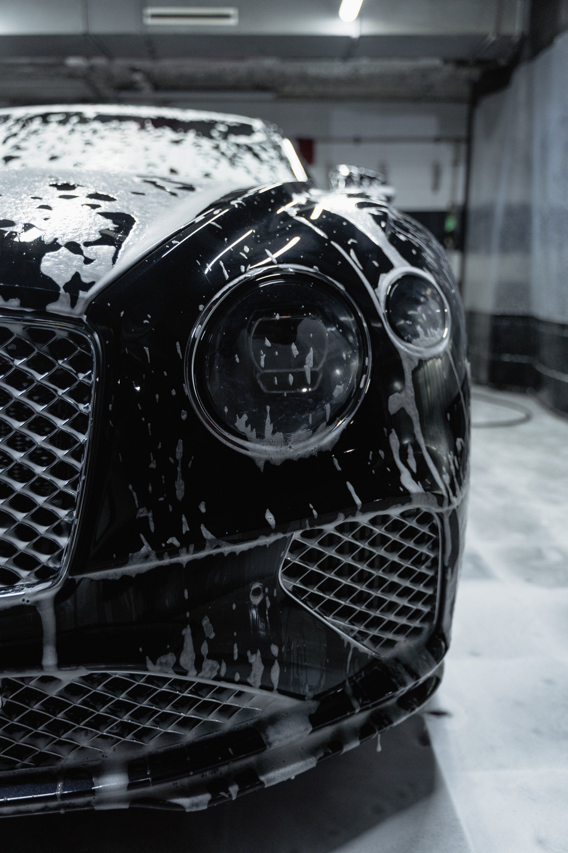 De voorkant van een zwarte Bentley, bedekt met zeepschuim, wordt gewassen in een garage.