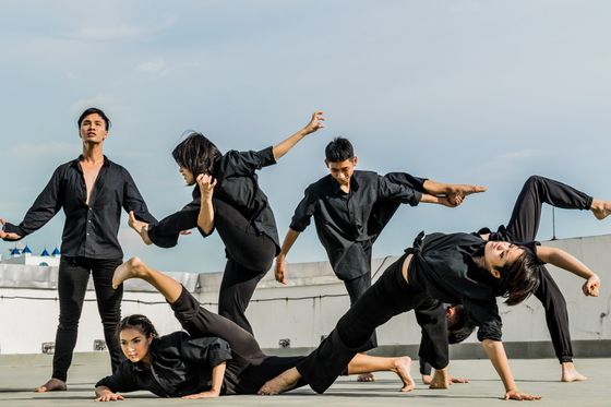 Dancers in black clothing perform a modern dance routine on a rooftop under a blue sky.