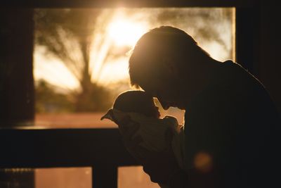 A man is holding a baby in his arms in front of a window while the sun is setting. 
